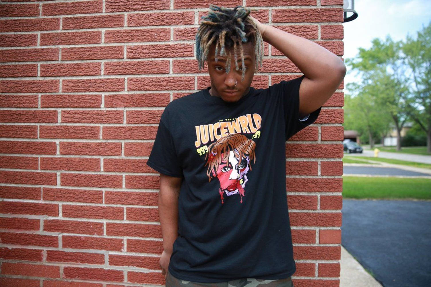 有彩色辮子的年輕人靠在磚牆上，穿著Juice WRLD T恤。 — Juice Wrld 壁紙