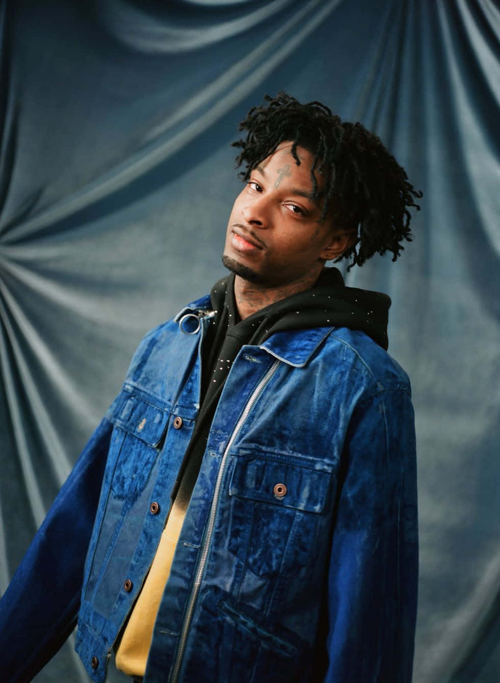 21 Savage 桌布 壁紙 — 一個有捲髮和面部刺青的男人，在藍色垂簾背景前，穿著藍色牛仔外套，內搭黑色連帽衫。