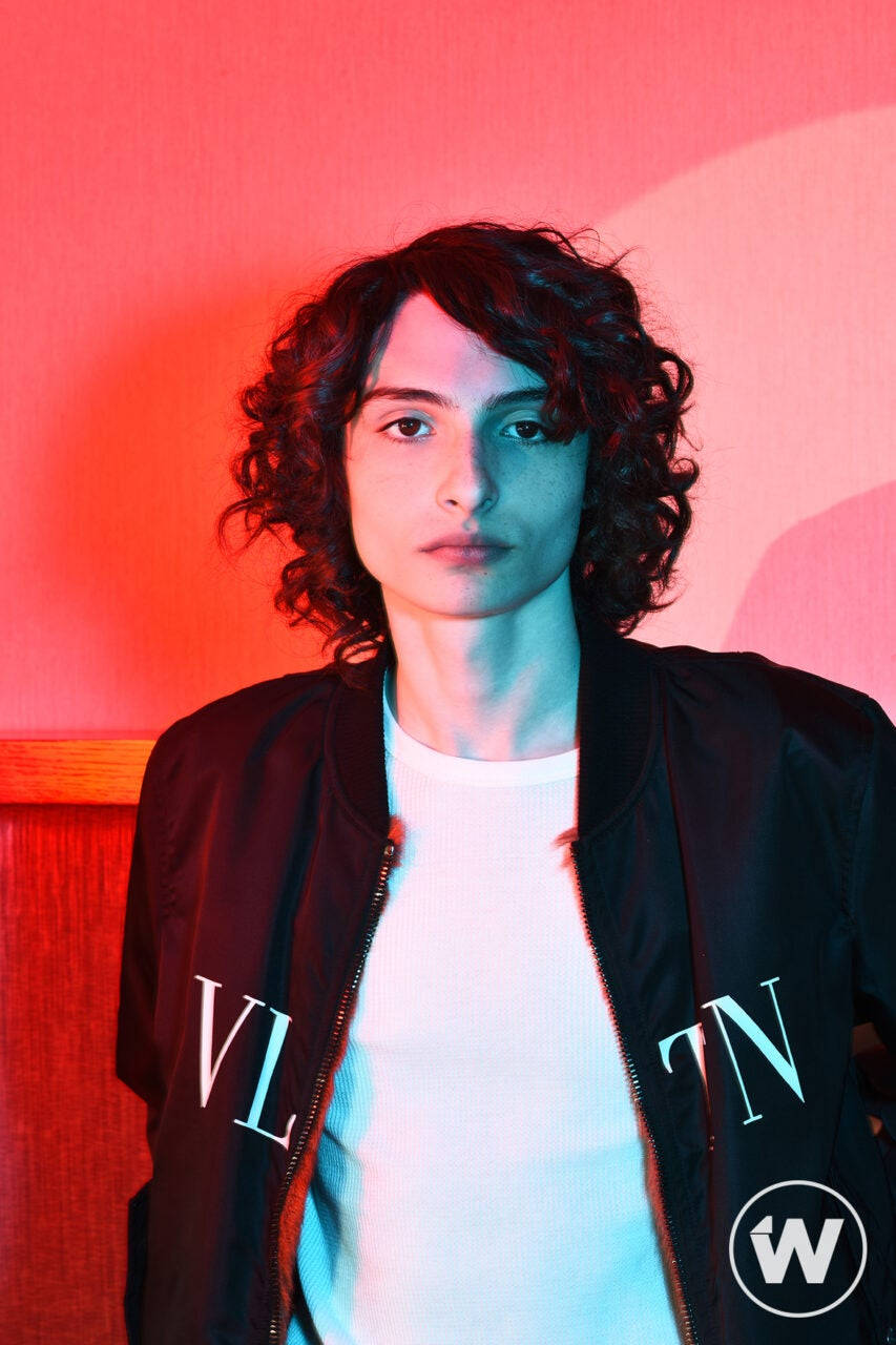 finn wolfhard 桌布 壁紙 — 一個有捲髮的人，在粉紅色牆壁前，紅色和藍色霓虹燈光下穿著黑色轟炸機外套。