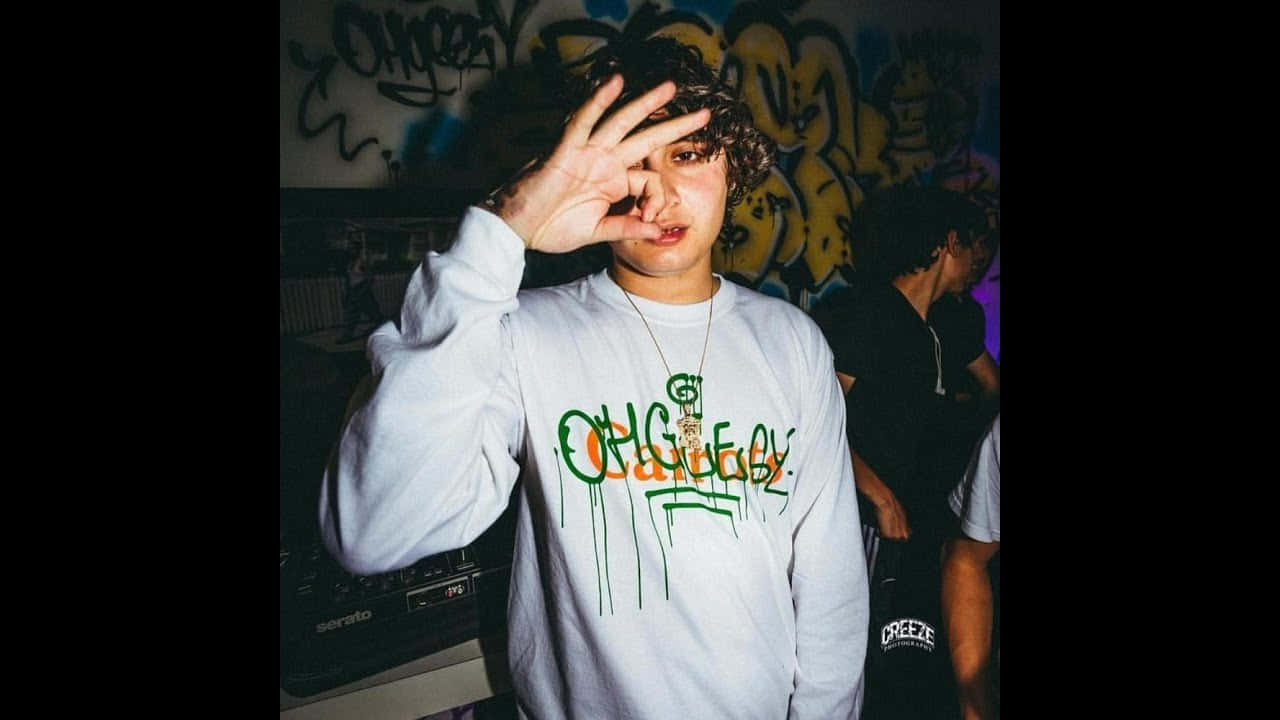 Shoreline Mafia 桌布 壁紙 — 一個有捲髮的年輕人在一個昏暗的城市環境中，在一個佈滿塗鴉的牆前面做了一個眼神動作。