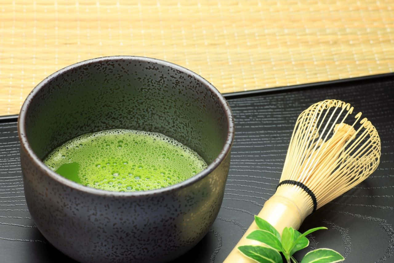 Matcha 桌布 壁紙 — 一個有斑點的陶瓷碗盛載著泡沫狀的抹茶，旁邊有竹筅和新鮮茶葉，放在黑色托盤上。