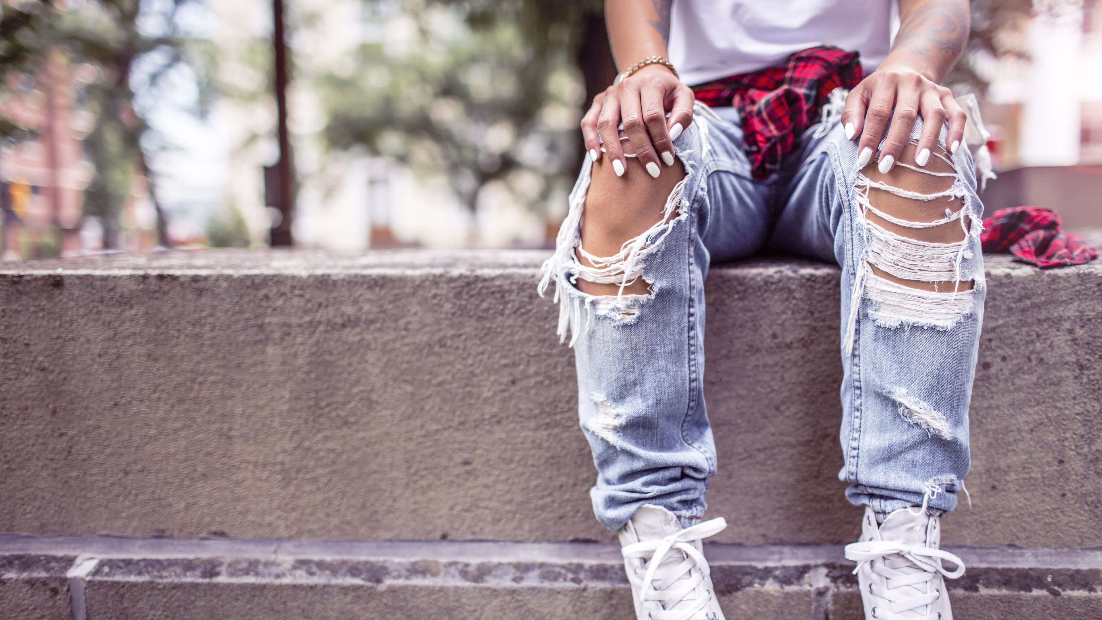 Jeans 桌布 壁紙 — 一個有白色指甲的人坐在混凝土台階上，穿著破洞牛仔褲和白色運動鞋，腰間繫著一件紅色格紋襯衫。