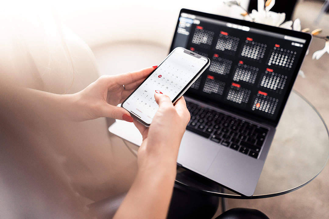 Valorant iPhone 壁紙 桌布 壁紙 — 一個有紅色指甲的人使用智慧型手機日曆應用程式，而一台顯示相同日曆的筆記型電腦放在玻璃桌上。