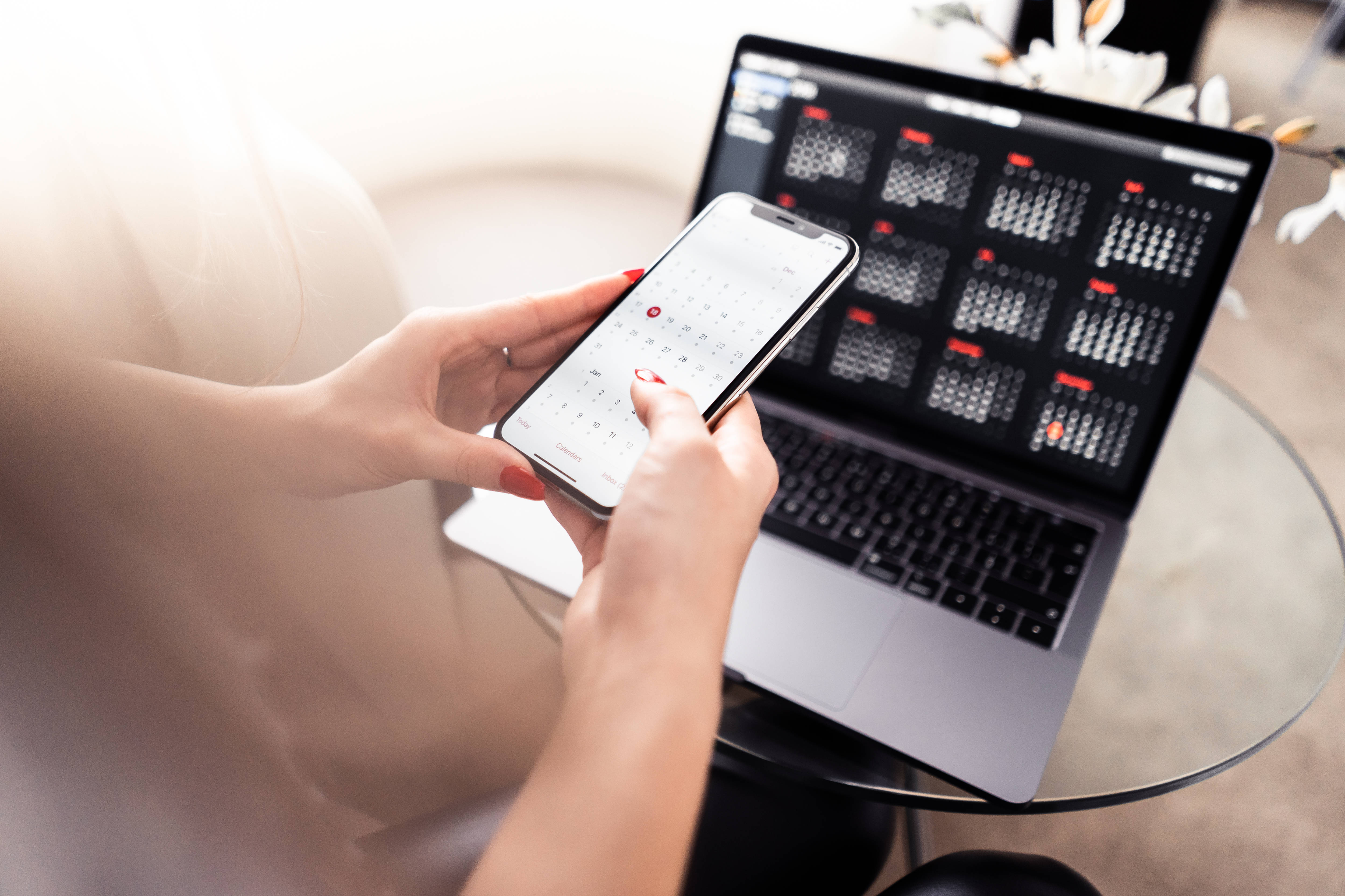 Valorant iPhone 壁紙 桌布 壁紙 — 一個有紅色指甲的人正在使用智慧型手機日曆應用程式，而筆記型電腦在玻璃桌上顯示一個匹配的數位日曆。
