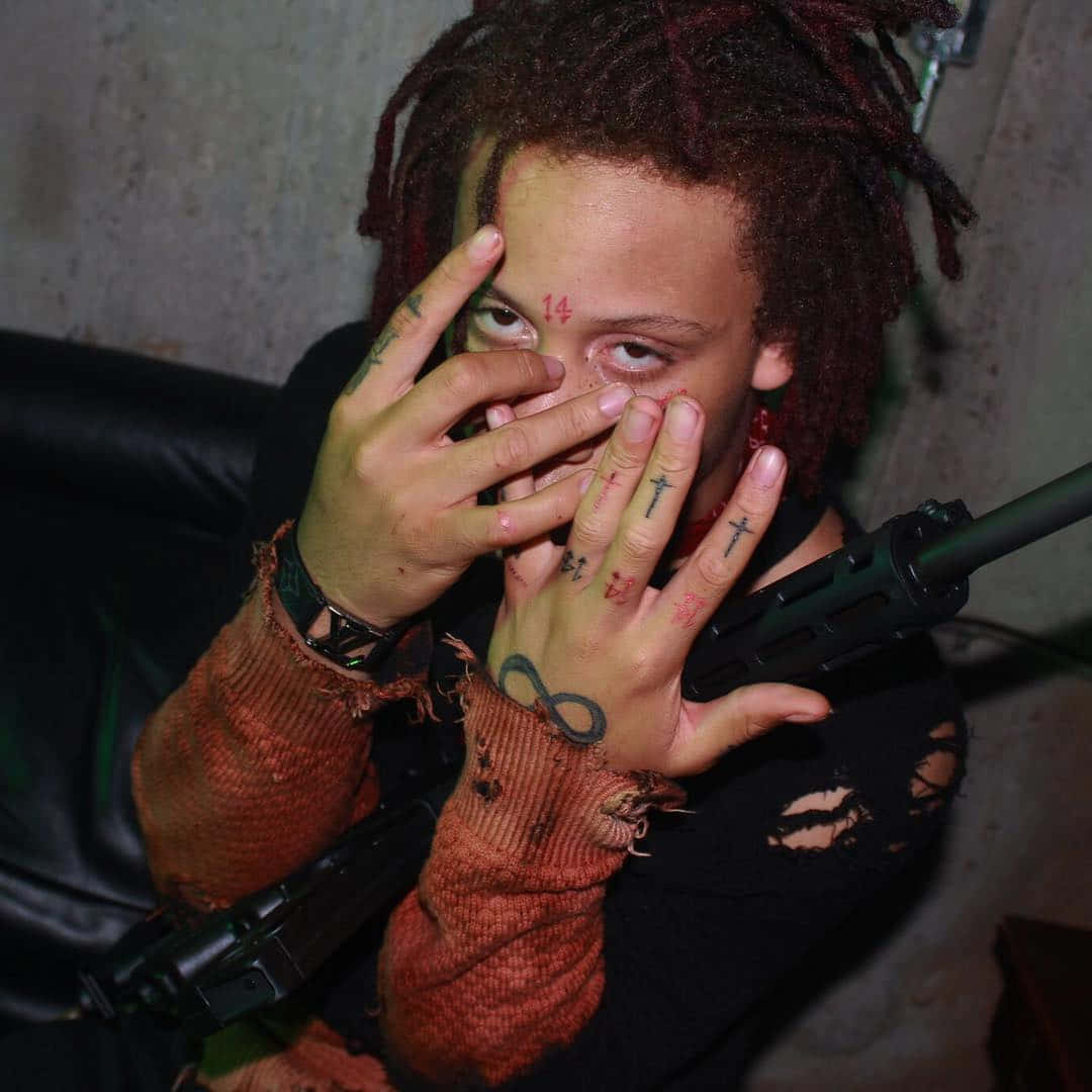 Trippie Redd 桌布 壁紙 — 一個有紅色辮子和刺青的人，在昏暗的房間裡用雙手遮住臉，同時拿著步槍。