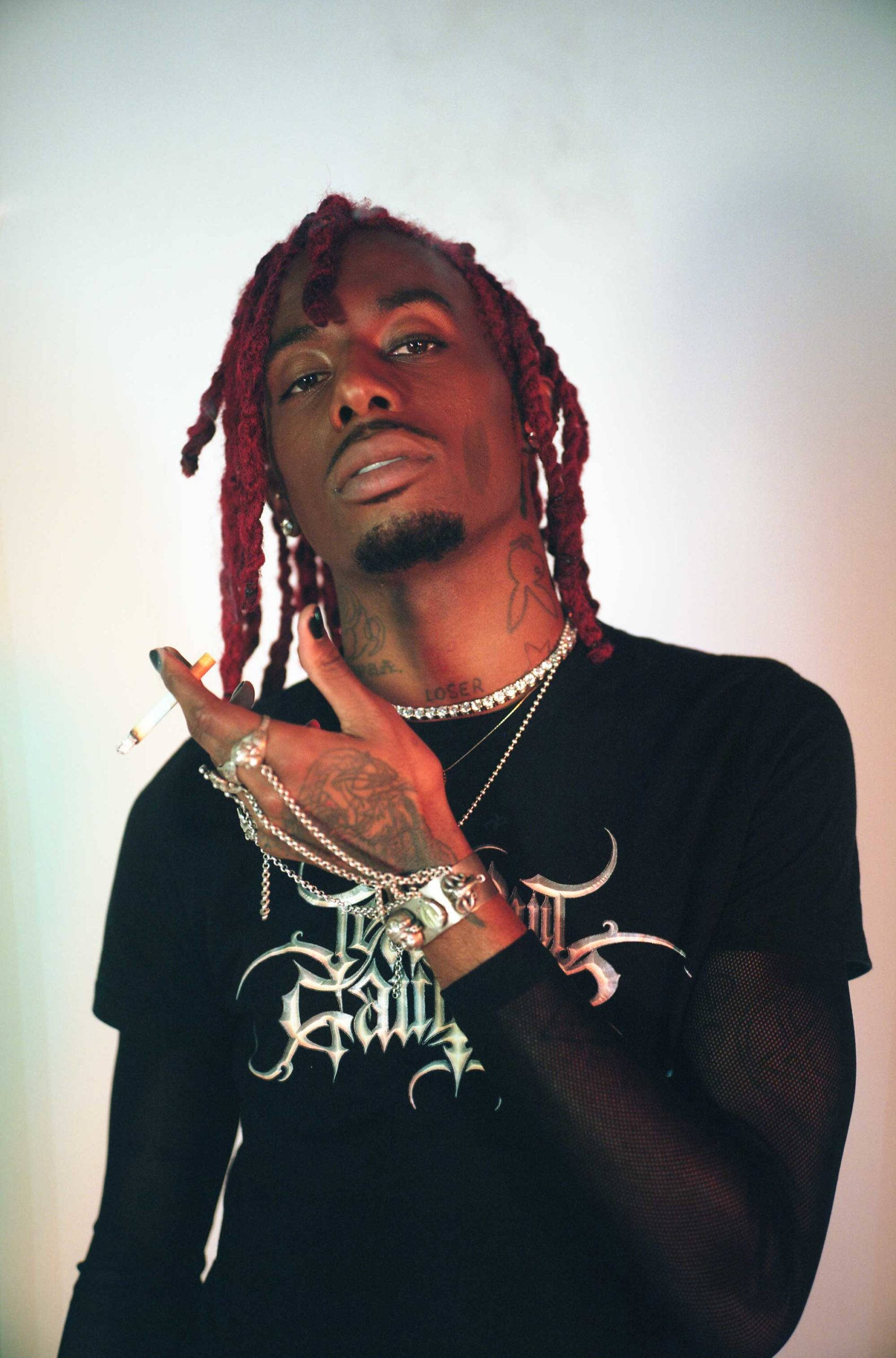 Carti 桌布 壁紙 — 一個有紅色辮子頭和刺青的男人，戴著多層銀鏈和黑色網眼衫，在柔和漸變背景前拿著一支煙。