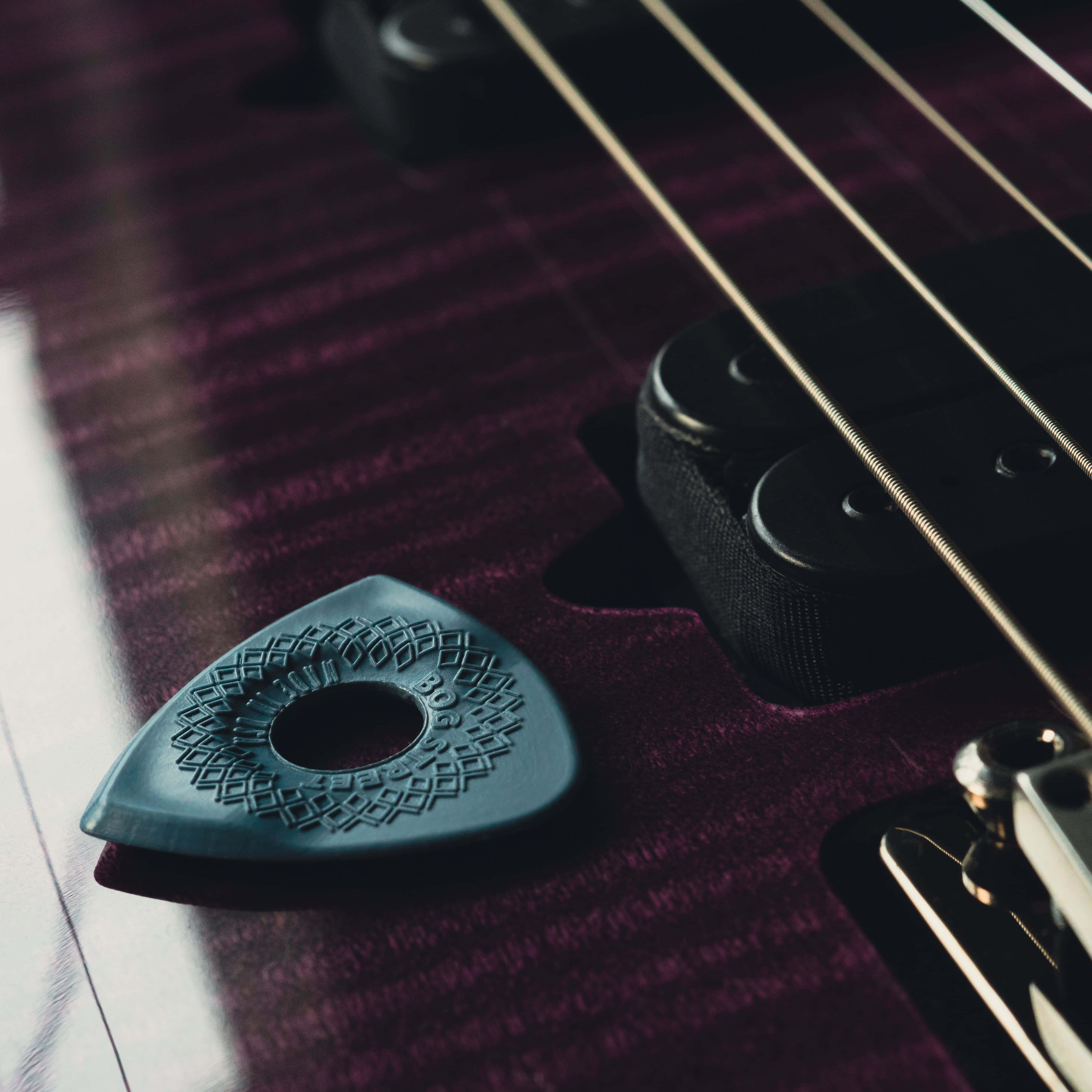Guitar Aesthetic 桌布 壁紙 — 一個有紋理的藍色吉他撥片特寫，放在深紫色木吉他琴身上，靠近金屬弦線和拾音器。