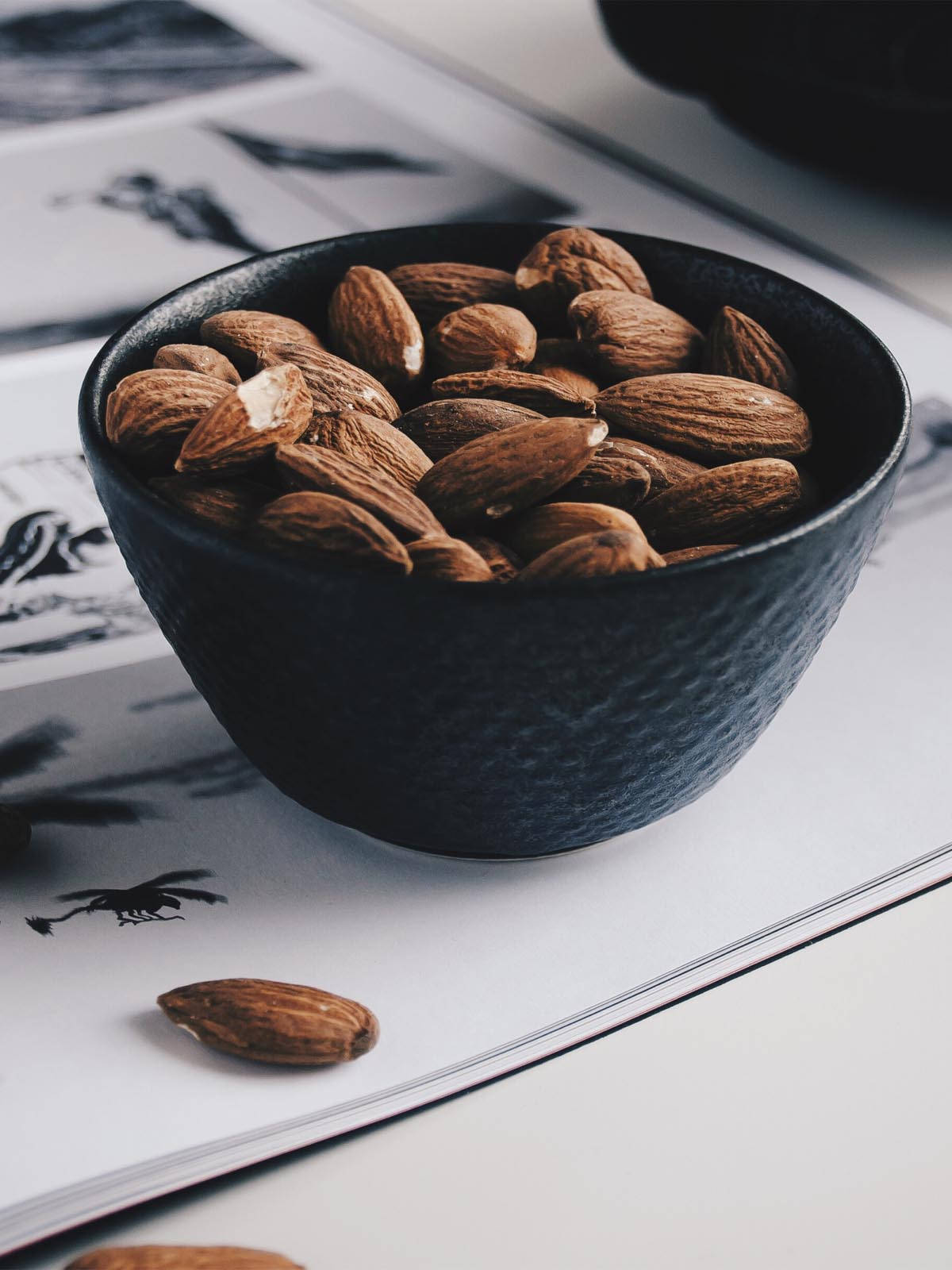Almond 桌布 壁紙 — 一個有紋理的黑色碗裝滿了杏仁，放在雜誌頁面上，附近散落著一顆杏仁。