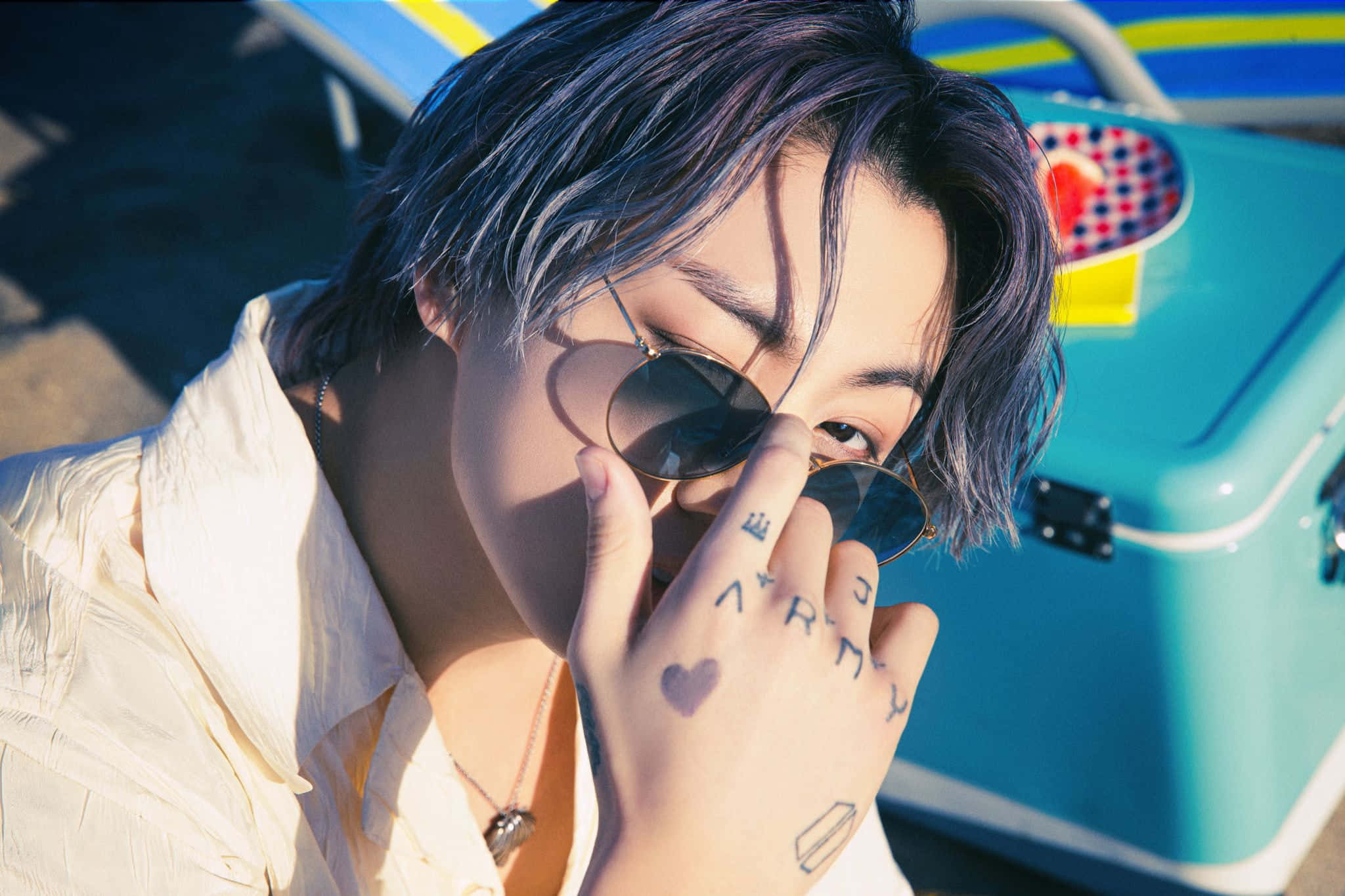 Jungkook 桌布 壁紙 — 一個有紫色頭髮和太陽眼鏡的人在明亮陽光下調整眼鏡，同時在藍色冷卻器附近展示手上的刺青。