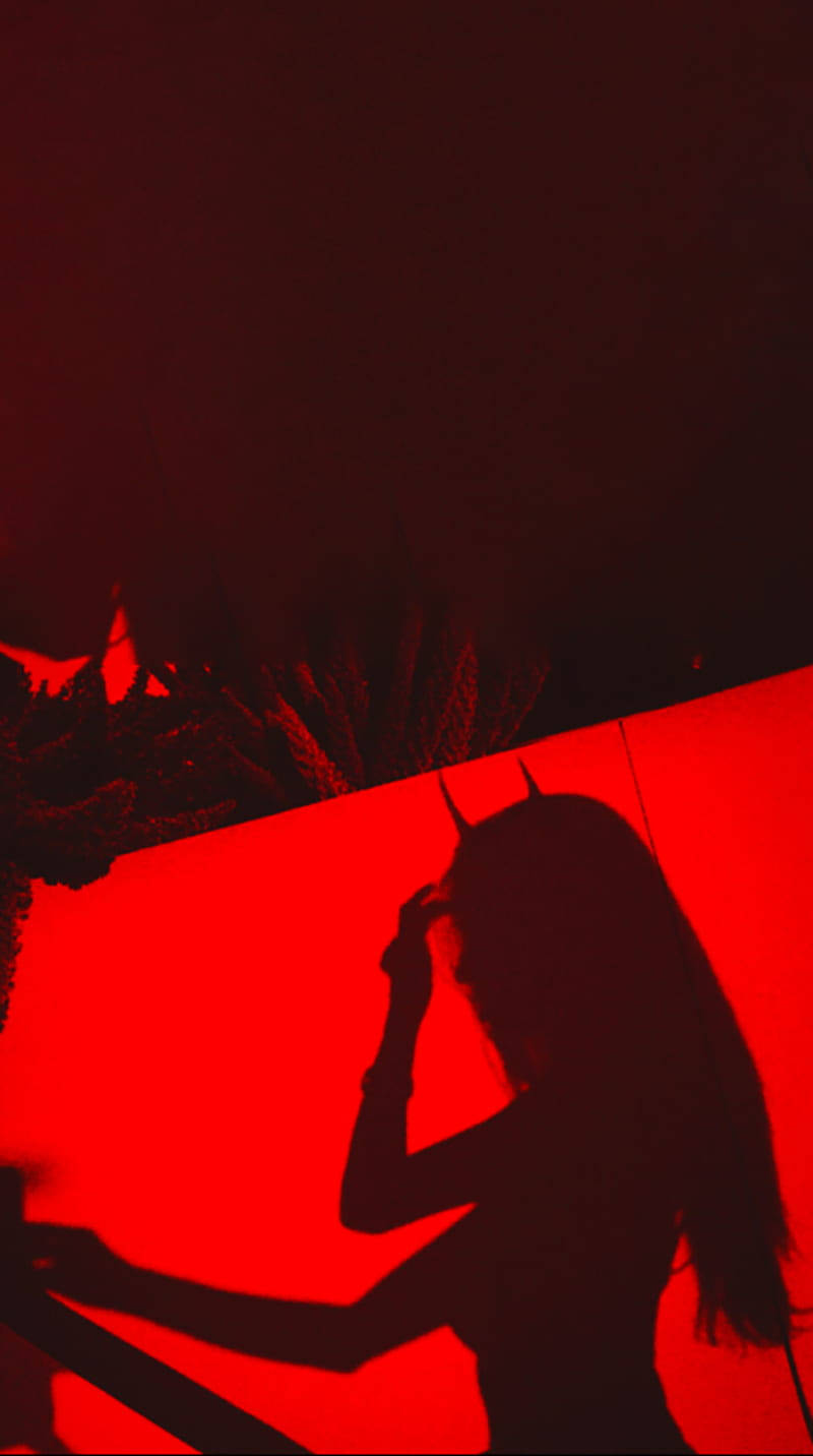 Sad Demon Girl 桌布 壁紙 — 一個有角的人物在鮮紅色背景和深色樹葉前調整頭飾的剪影。