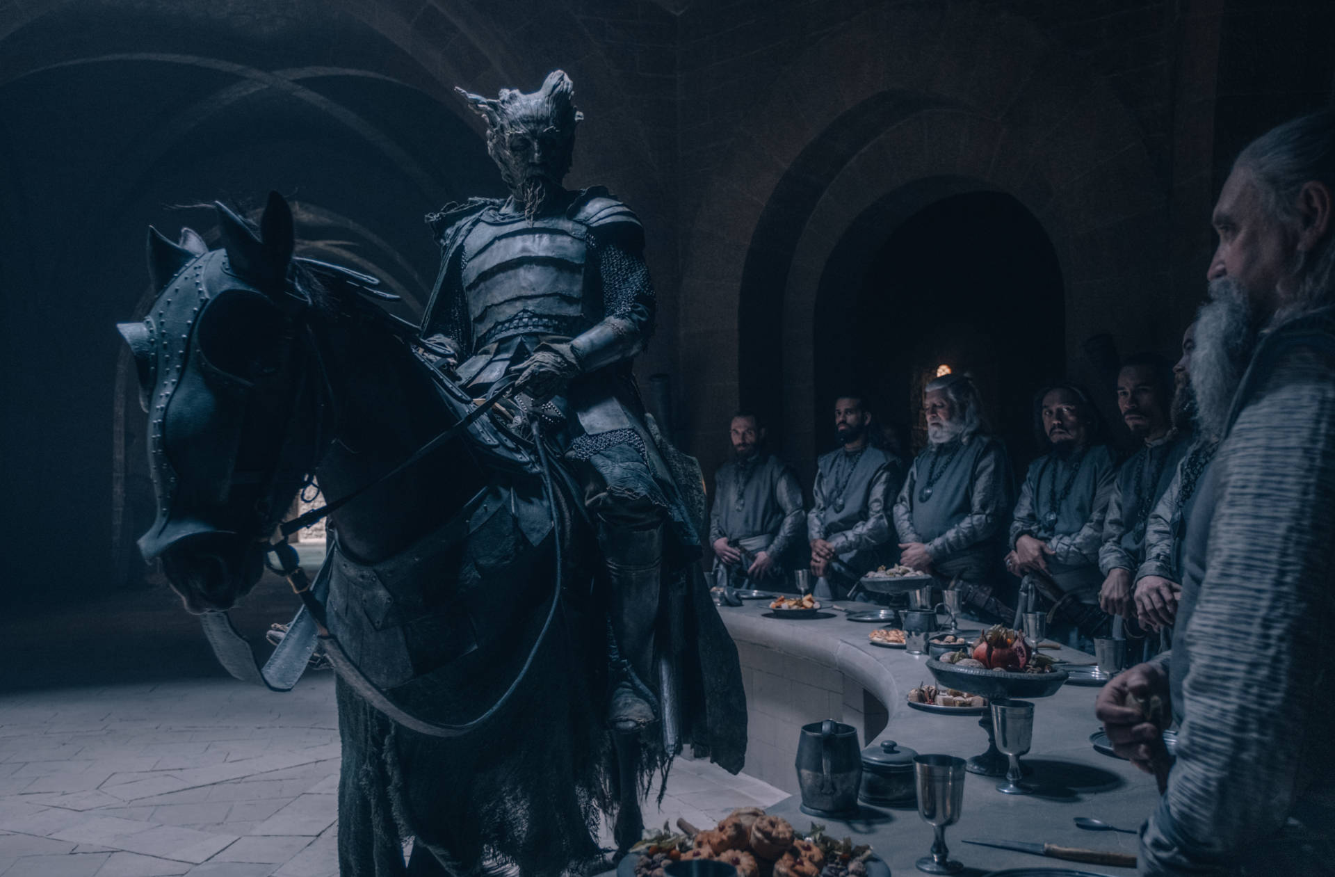 The Green Knight 桌布 壁紙 — 一個有角的裝甲人物騎著黑馬，威嚴地站在一個昏暗石廳中坐著的議會面前。