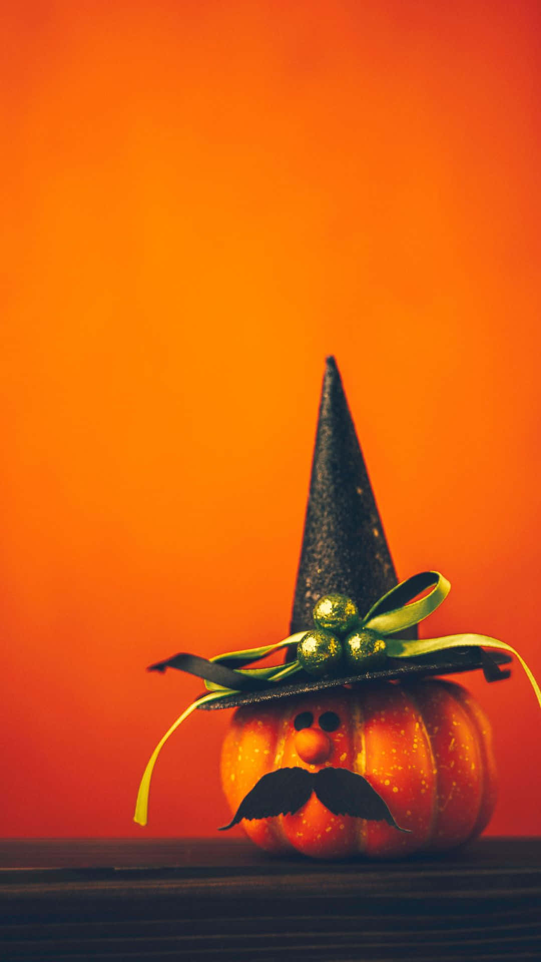 Cute Pumpkin 桌布 壁紙 — 一個有趣的橙色南瓜，戴著黑色巫師帽和鬍子，放在木質表面上，背景是溫暖的橙色漸變。