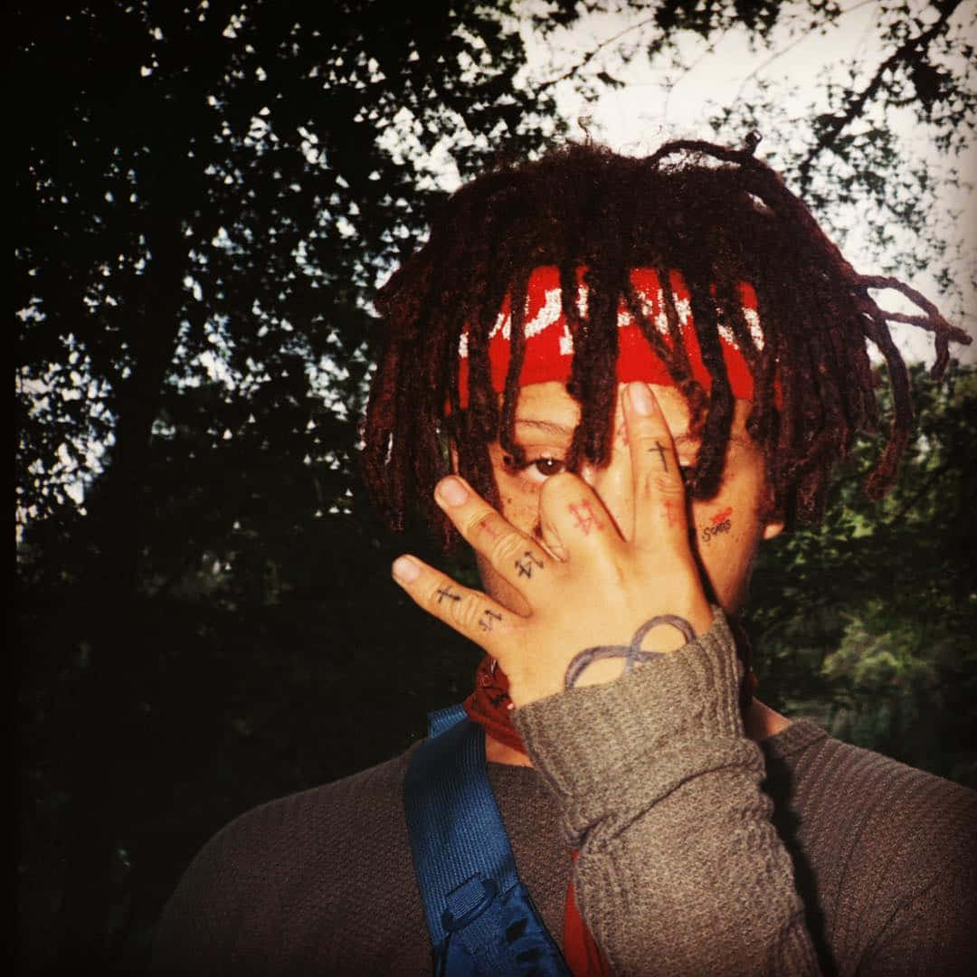 Trippie Redd 桌布 壁紙 — 一個有辮子頭和刺青的人站在樹林地區，用手遮住半邊臉。