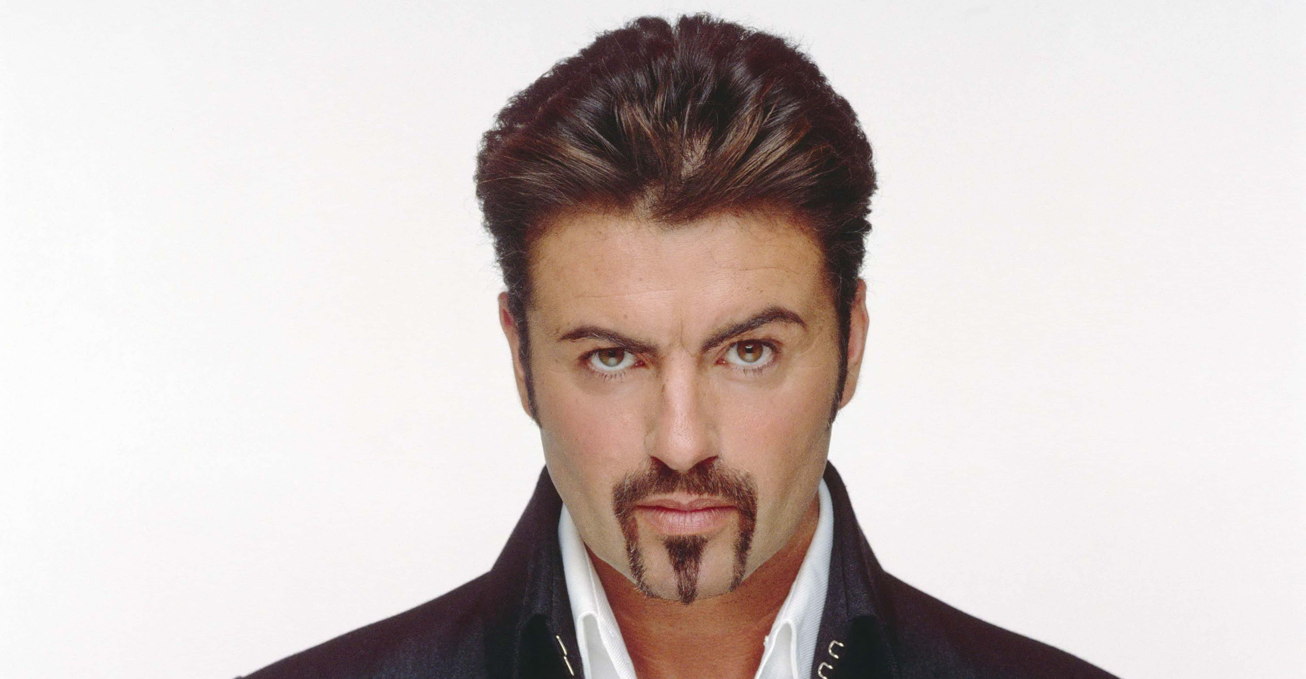 George Michael 桌布 壁紙 — 一個有造型深色頭髮和山羊鬍的男人，穿著深色外套內搭白襯衫，在純白背景前凝視。