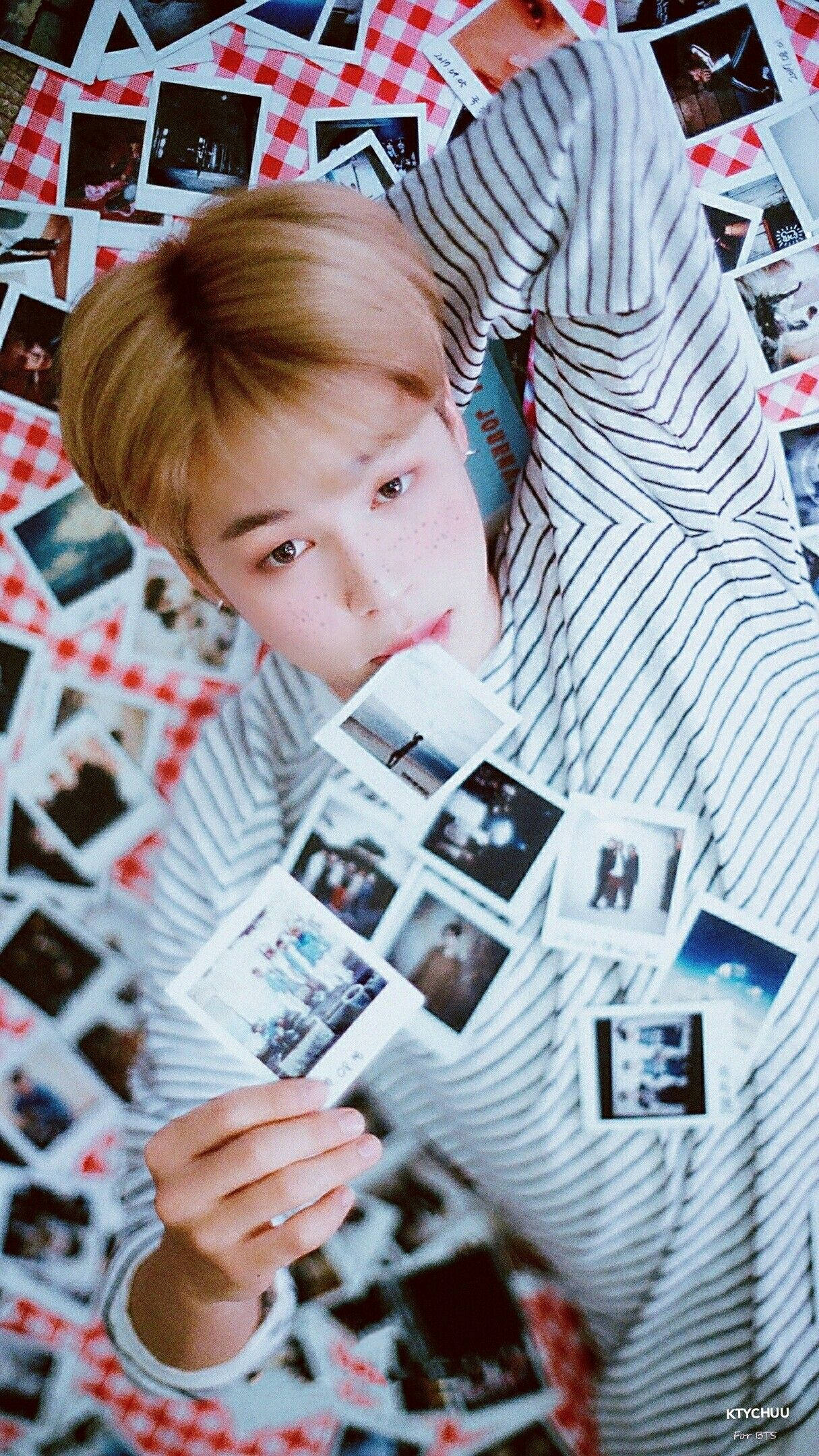 BTS 桌布 壁紙 — 一個有雀斑的年輕男子穿著條紋襯衫，躺在紅色格紋表面上，周圍散落著拍立得照片。