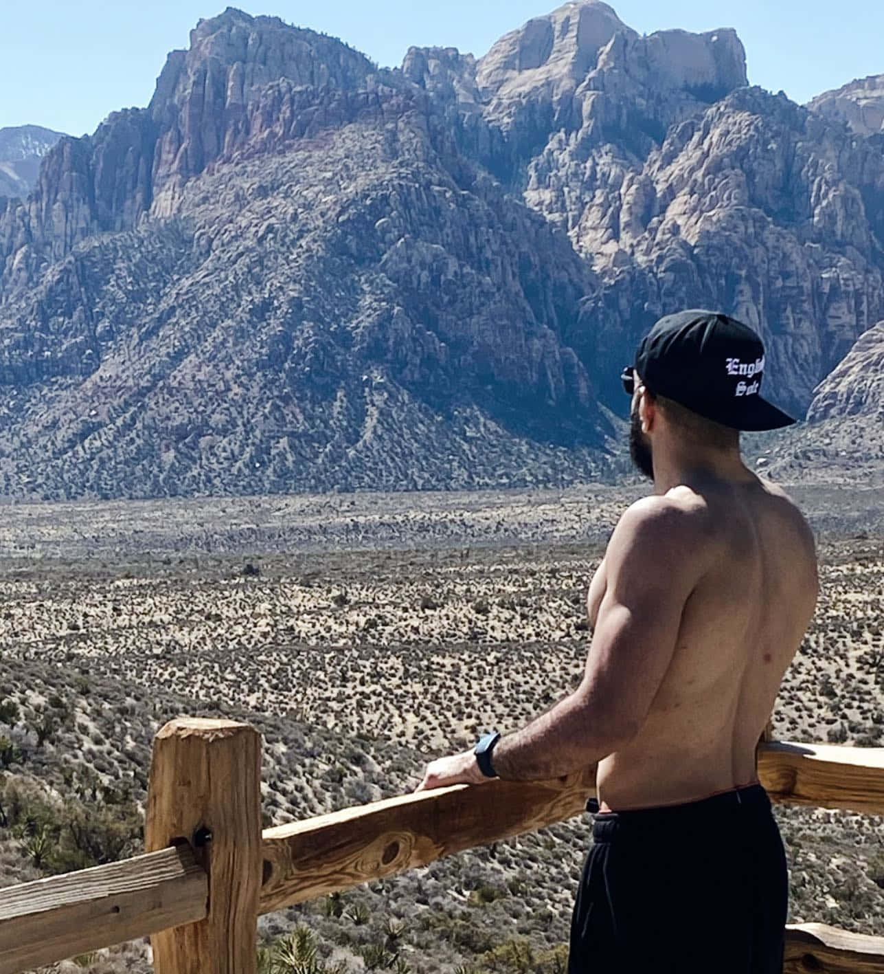 Vacation 桌布 壁紙 — 一個有鬍鬚的赤裸上身男人站在木欄杆旁，凝視著晴朗藍天下高聳的岩石山脈。