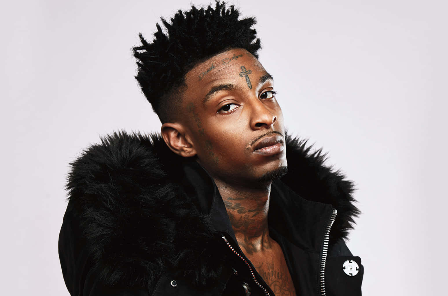 21 Savage 桌布 壁紙 — 一個有黑色尖刺頭髮和面部刺青的男人，在純白色背景前穿著黑色毛領夾克。