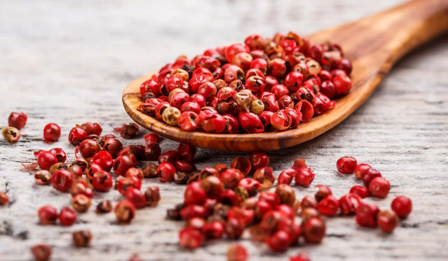 Pink Peppercorn 桌布 壁紙 — 一個木勺盛滿鮮紅的胡椒粒，放在經風化的灰色木質表面上，周圍散落著種子。