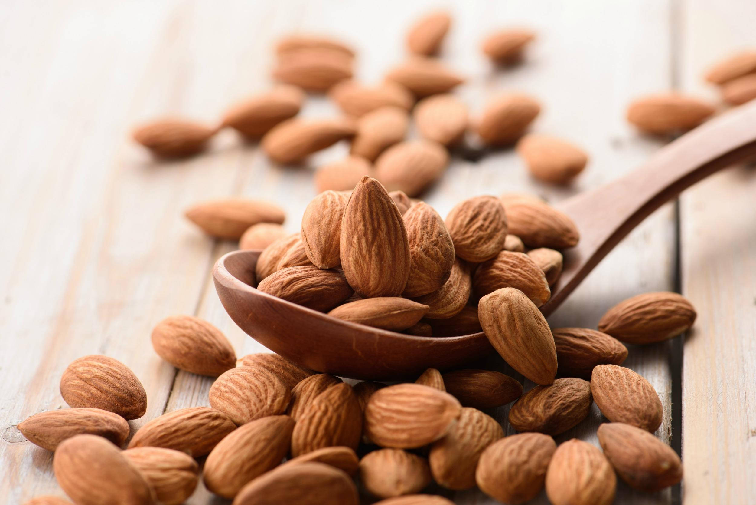 Almond 桌布 壁紙 — 一個木勺裝滿杏仁，溢出到淺色木質表面上，景深較淺。