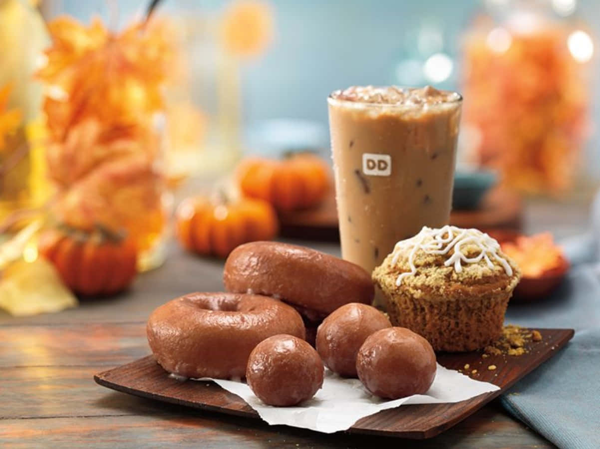 Dunkin Donuts 桌布 壁紙 — 一個木托盤上放有釉面甜甜圈、巧克力松露和南瓜香料馬芬，旁邊是一杯冰咖啡，背景中有秋葉和南瓜，呈現柔和焦點。