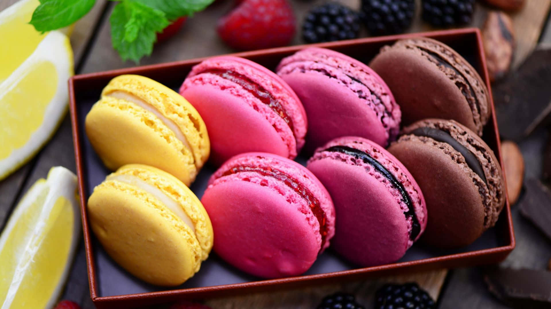 Desserts 桌布 壁紙 — 一個木托盤上放著色彩繽紛的馬卡龍，內餡有檸檬、覆盆子和巧克力，周圍環繞著新鮮漿果和薄荷。