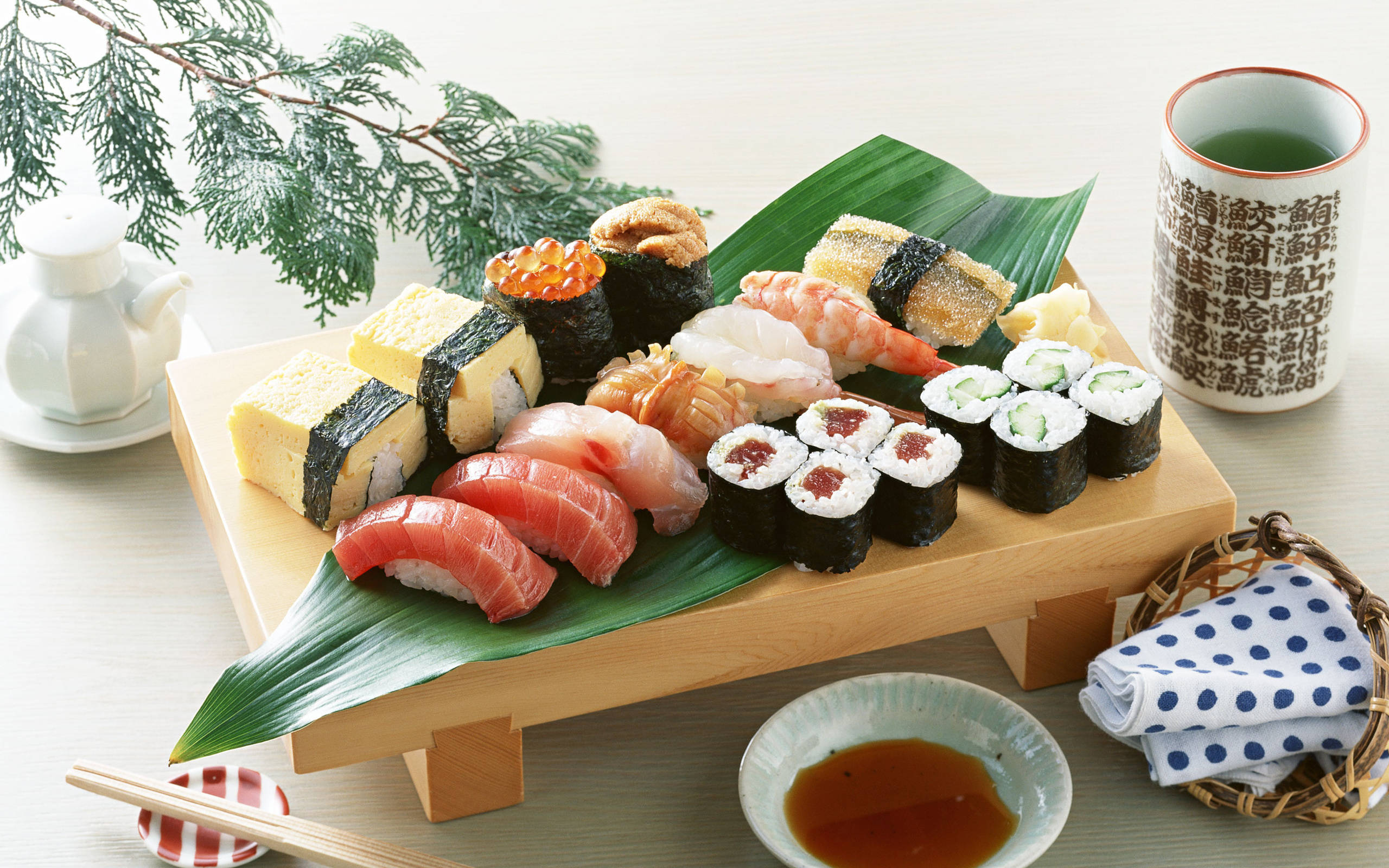 Sushi 桌布 壁紙 — 一個木托盤盛裝著各種色彩繽紛的壽司，擺放在綠葉上，旁邊有茶和筷子。