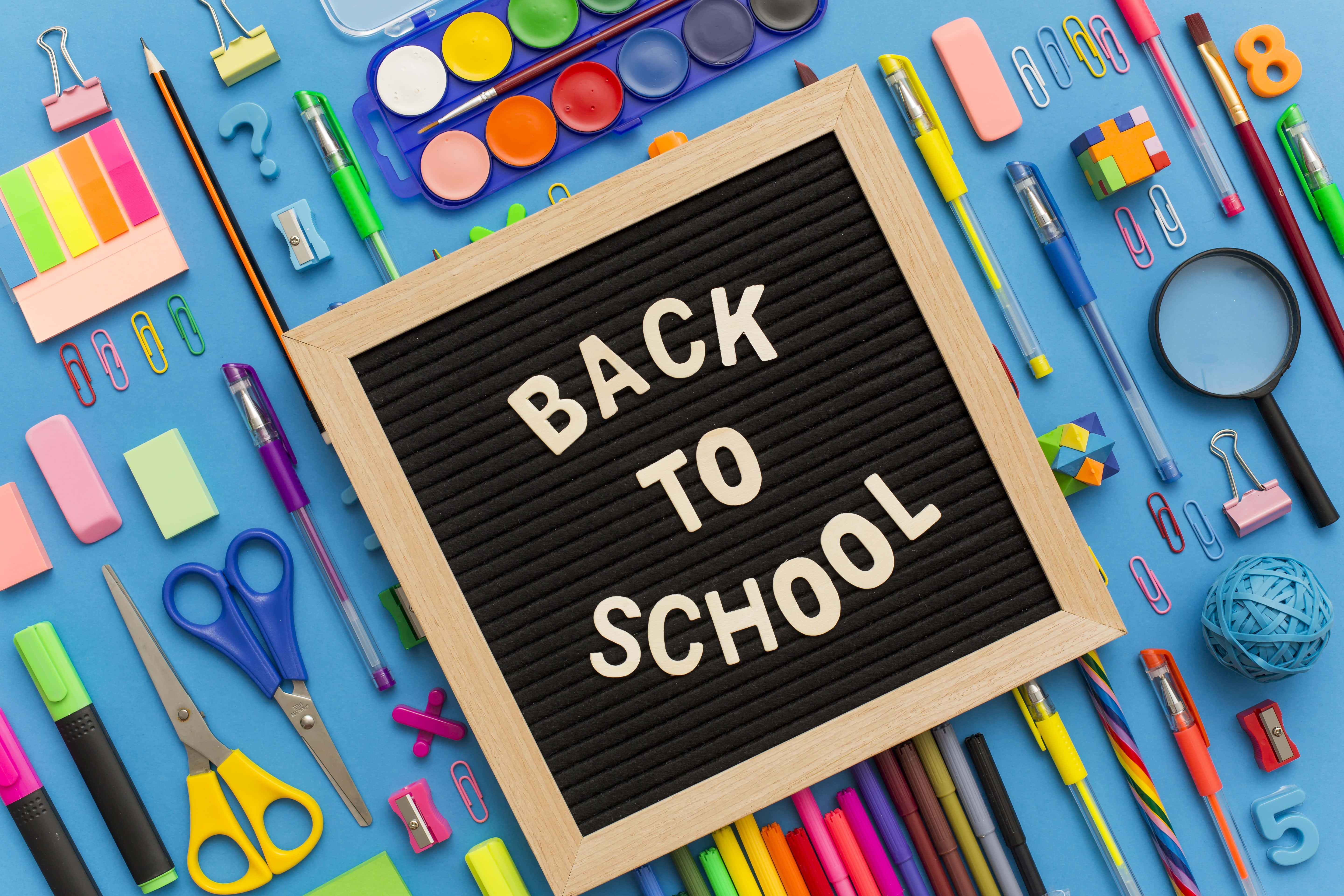 學校 桌布 壁紙 — 一個木框字母板，上面寫著「BACK TO SCHOOL」，周圍是色彩繽紛的文具，放在明亮的藍色表面上。