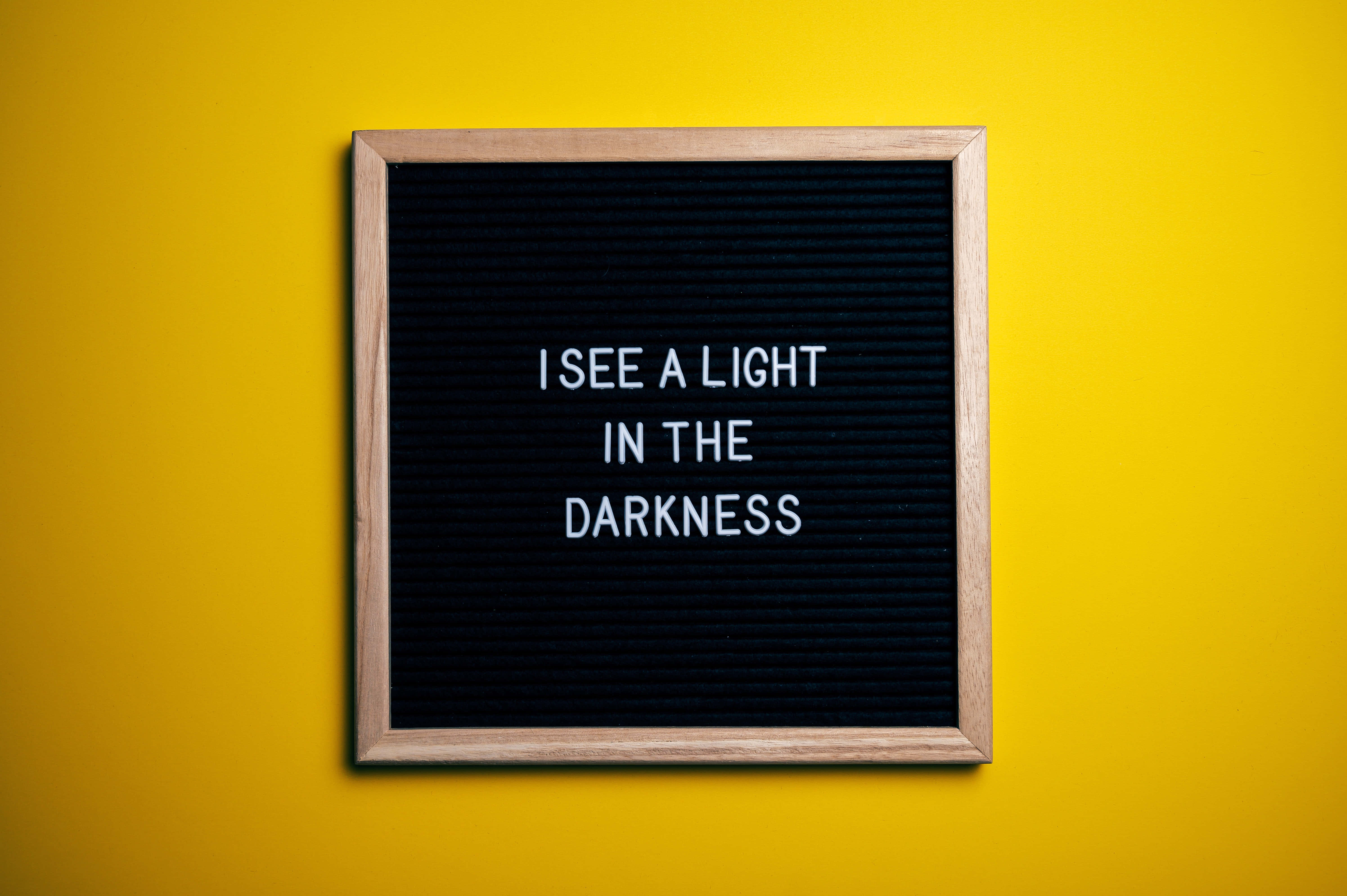 語錄 桌布 壁紙 — 一個木框字母板，白色字母寫著「I SEE A LIGHT IN THE DARKNESS」，背景是鮮黃色的牆壁。