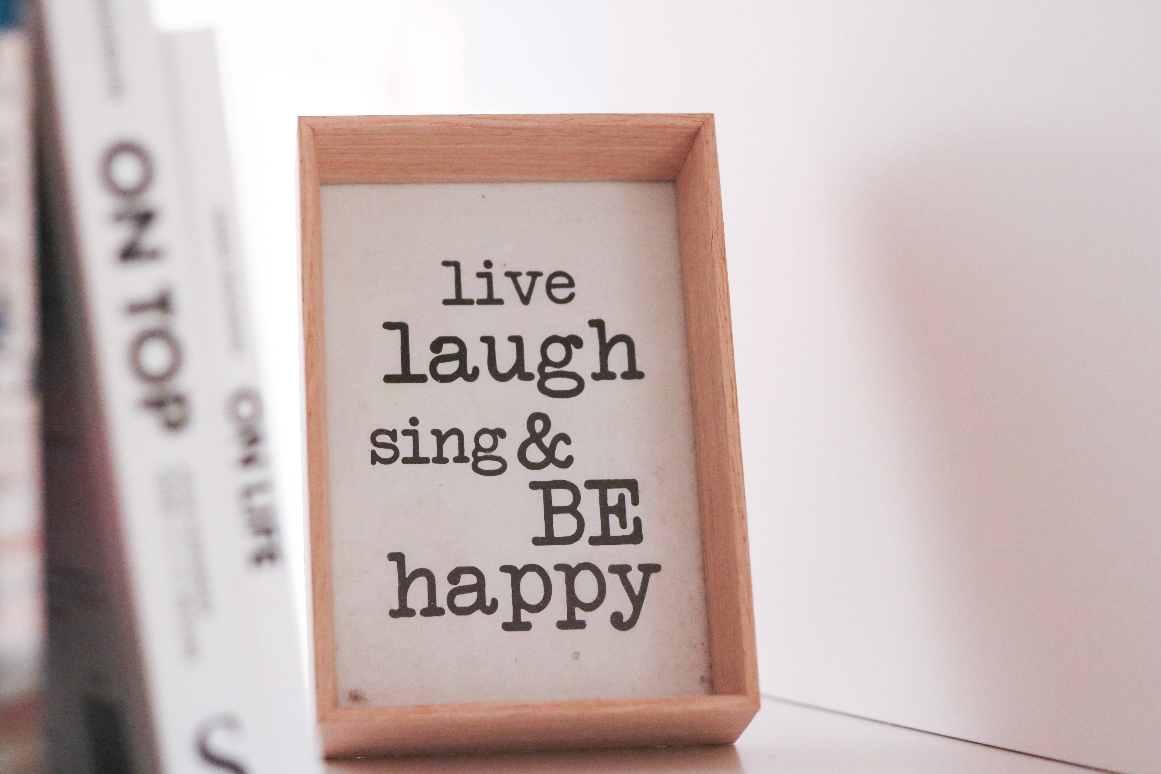 快樂 桌布 壁紙 — 一個木框標牌，上面寫著 live laugh sing & BE happy，放在書架上的書本旁邊。
