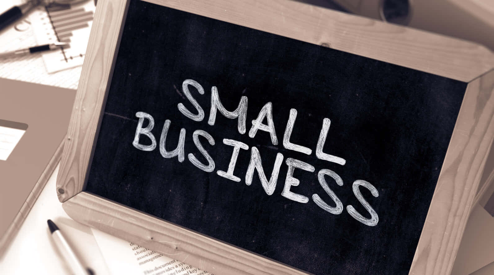 商業 桌布 壁紙 — 一個木框黑板，上面用白色粉筆寫著「SMALL BUSINESS」，放在一張被辦公用品包圍的桌子上。