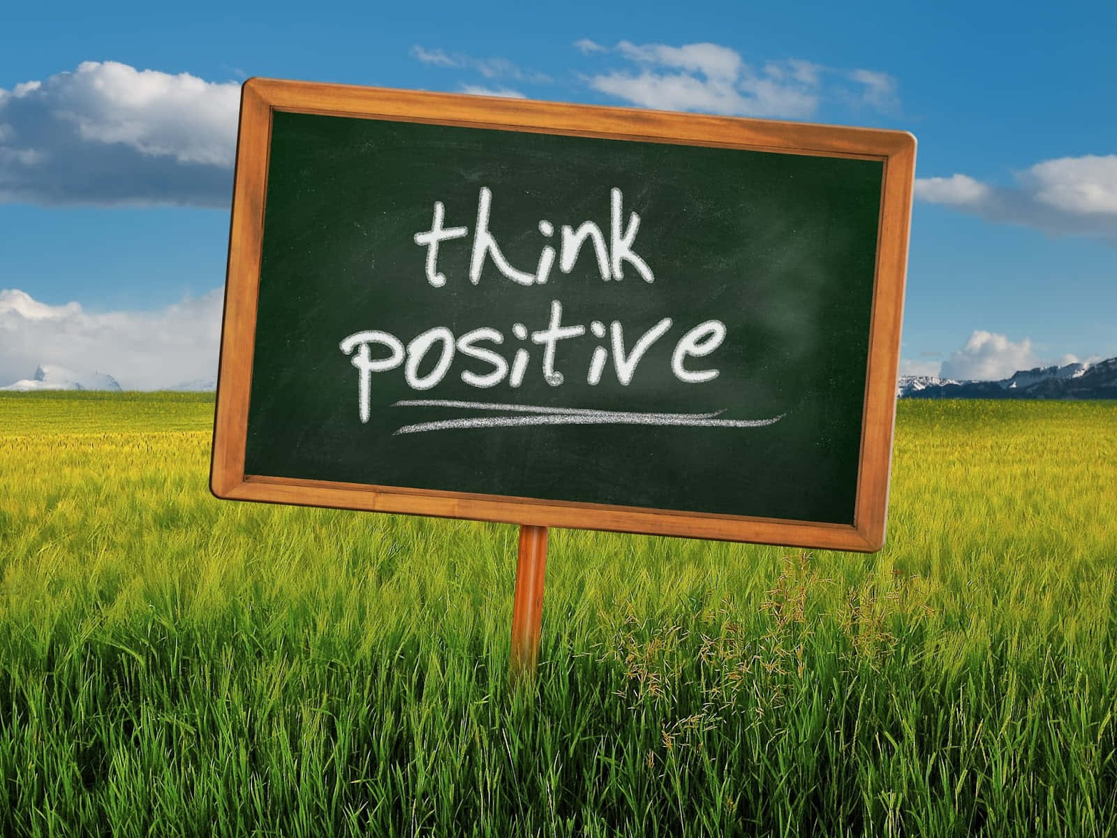 正向 桌布 壁紙 — 一個木框黑板在陽光明媚的田野中，寫著 think positive，背景是藍天和遠處的雪山。