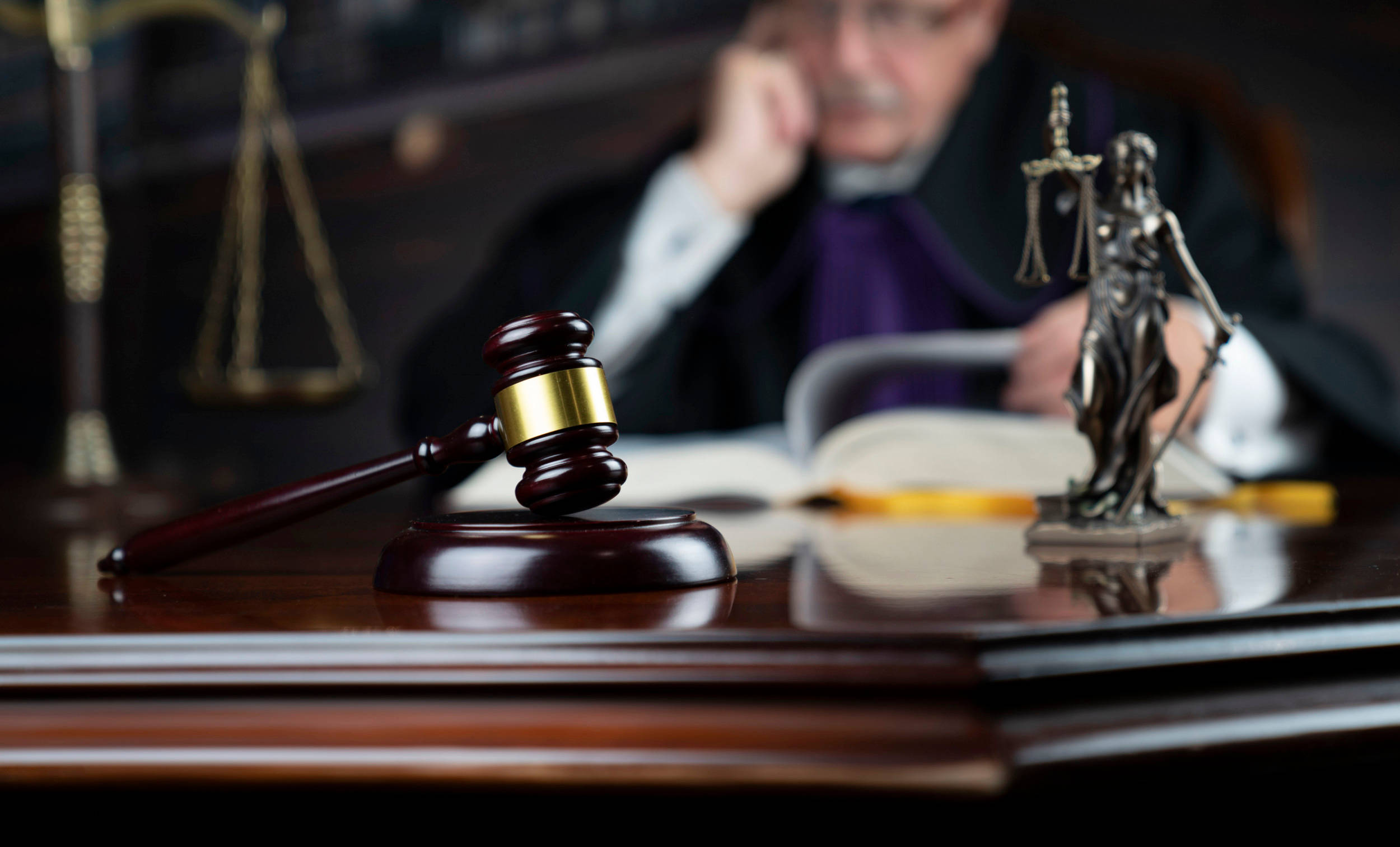 Lawyer 桌布 壁紙 — 一個木槌放在拋光桌上，旁邊有正義女神雕像和天平，背景有法官模糊的身影。