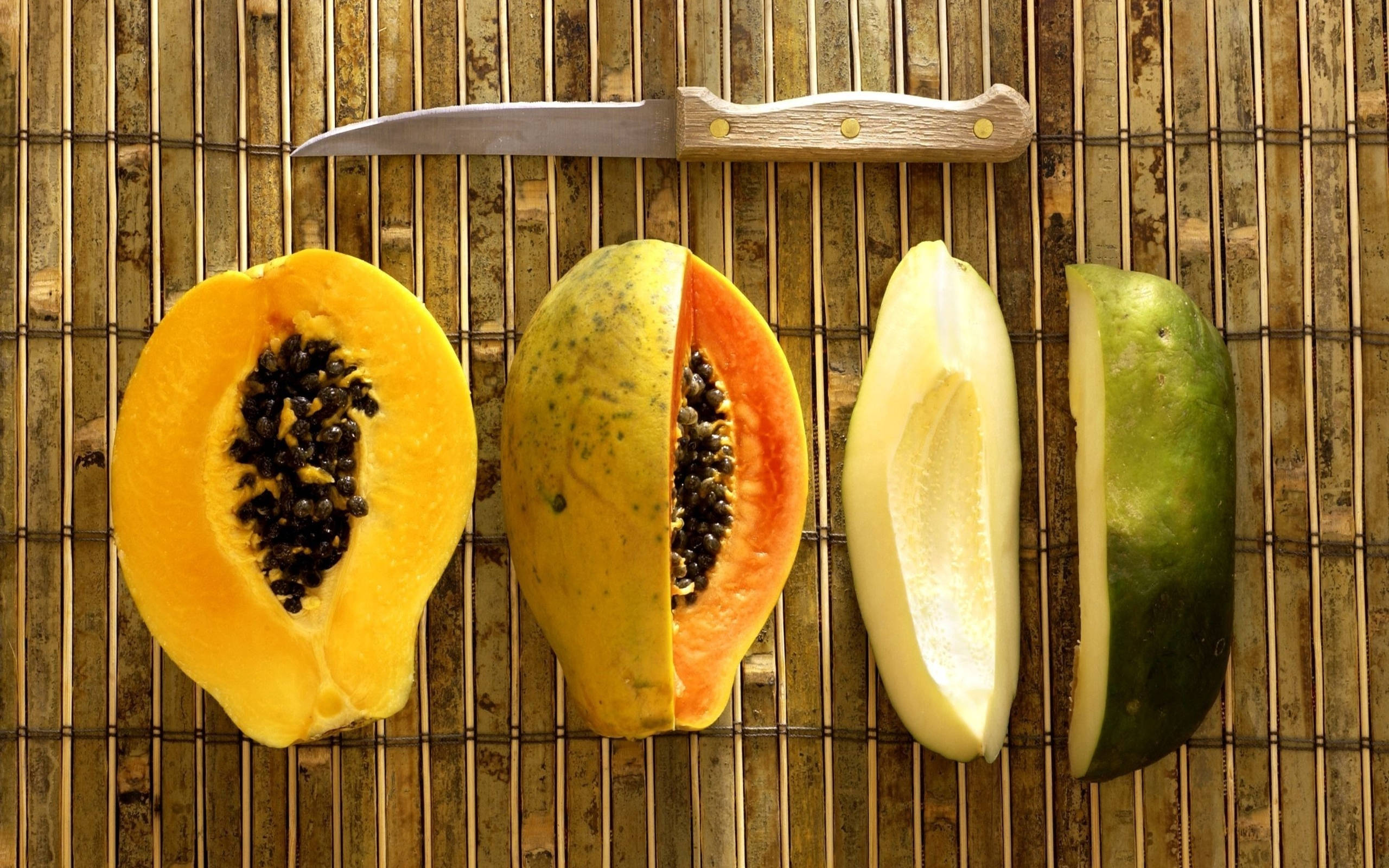 Fruits 桌布 壁紙 — 一個木瓜從完整到去皮的不同階段切片，露出多汁的橙色果肉和黑色種子，放在竹蓆上，旁邊有一把刀。