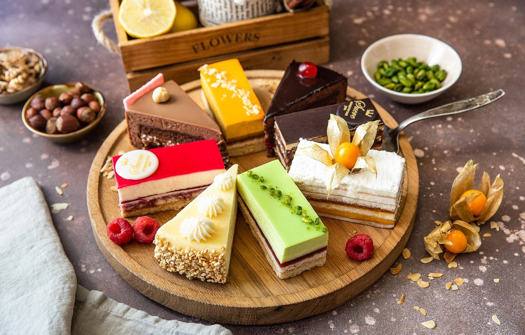 Bakery 桌布 壁紙 — 一個木盤上展示七塊色彩鮮豔的美食甜點切片，周圍環繞著新鮮漿果、堅果和裝飾豆莢，置於質樸的斑點表面上。
