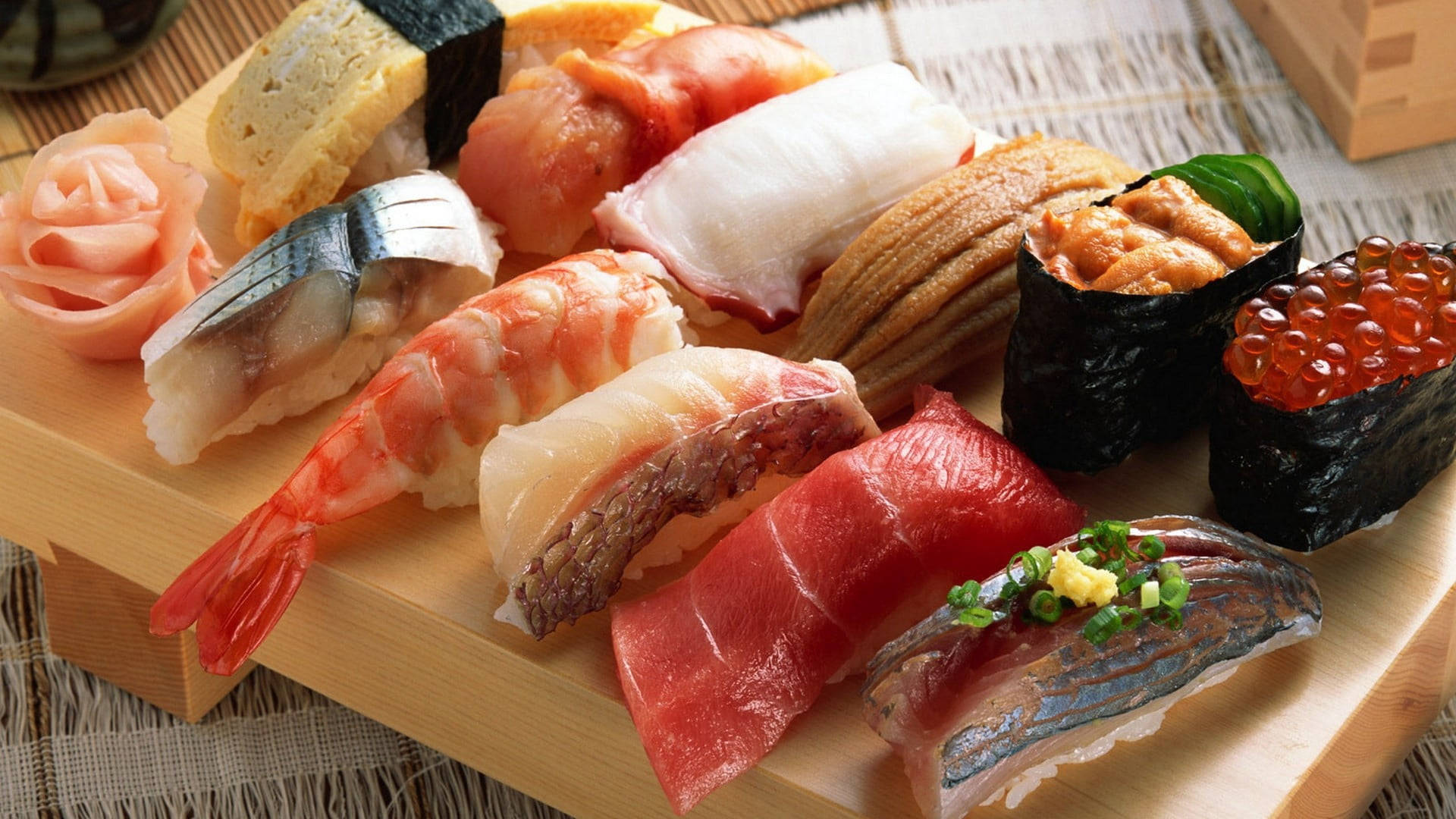 Sushi 桌布 壁紙 — 一個木盤上展示著各種色彩繽紛的壽司，包括鮪魚、蝦和鮭魚卵，整齊地排列在編織墊上。