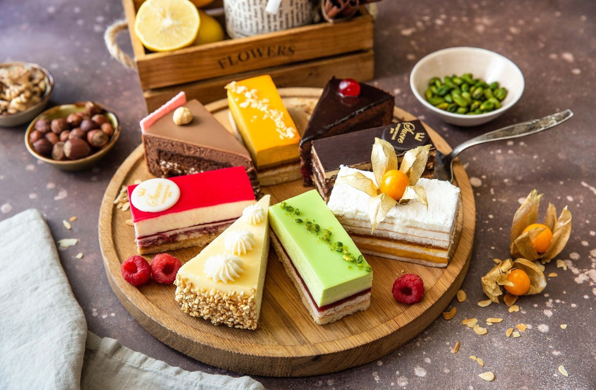 Pastry 桌布 壁紙 — 一個木盤上展示著色彩繽紛的美食蛋糕片，周圍環繞著新鮮漿果、堅果和裝飾元素，置於鄉村風格的桌面上。