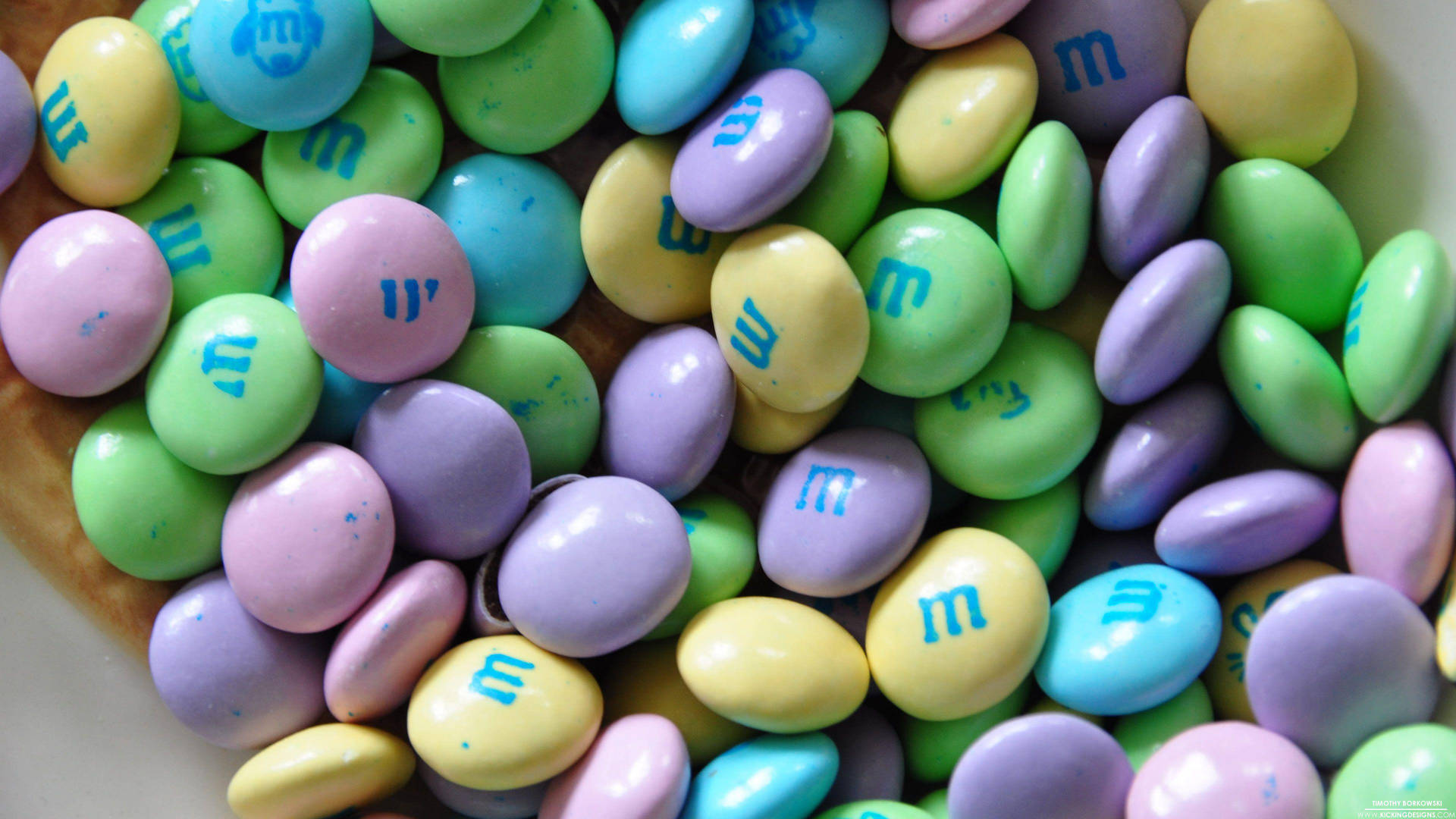 壁紙 — 一個木碗中散落著色彩繽紛的粉彩M&M's糖果，帶有藍色「m」標誌的特寫。