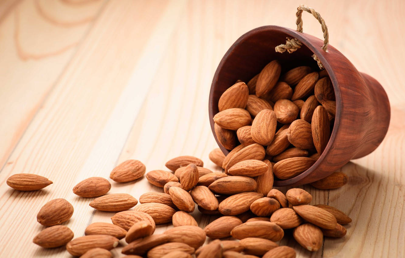Almond 桌布 壁紙 — 一個木碗傾倒，將杏仁灑在淺色木質表面上，光線溫暖自然。