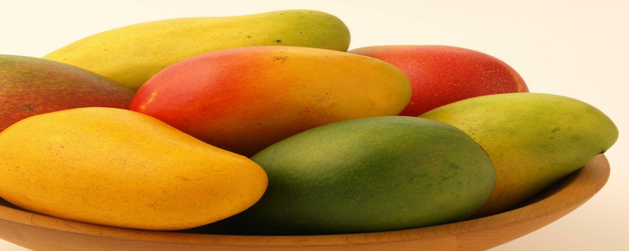 Mango 桌布 壁紙 — 一個木碗盛裝著色彩繽紛的成熟芒果，呈現鮮豔的黃色、紅色和綠色色調，背景為純白色。