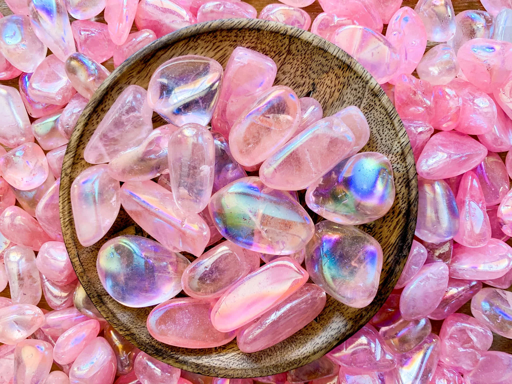 Pink Crystal Quartz 桌布 壁紙 — 一個木碗裝滿了虹彩粉紅石英石，周圍還有更多石頭放在木質表面上。