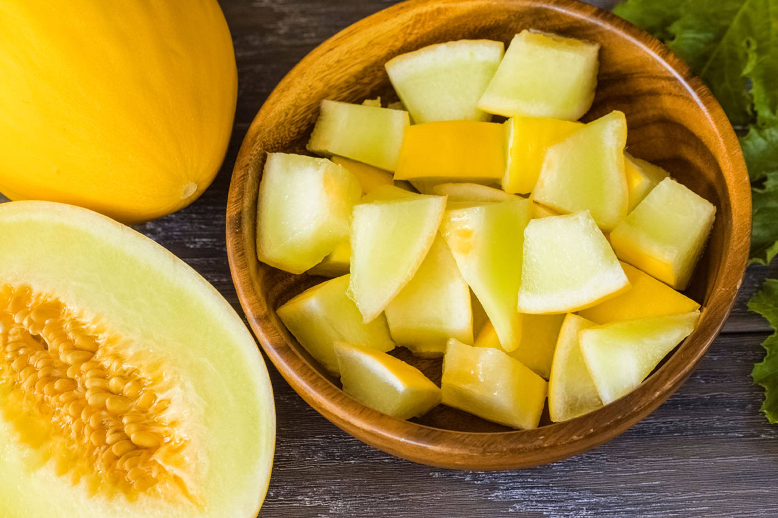 Fruit 桌布 壁紙 — 一個木碗裝滿多汁的黃色蜜瓜塊，旁邊放著半個蜜瓜和新鮮蔬菜，在深色木質表面上。