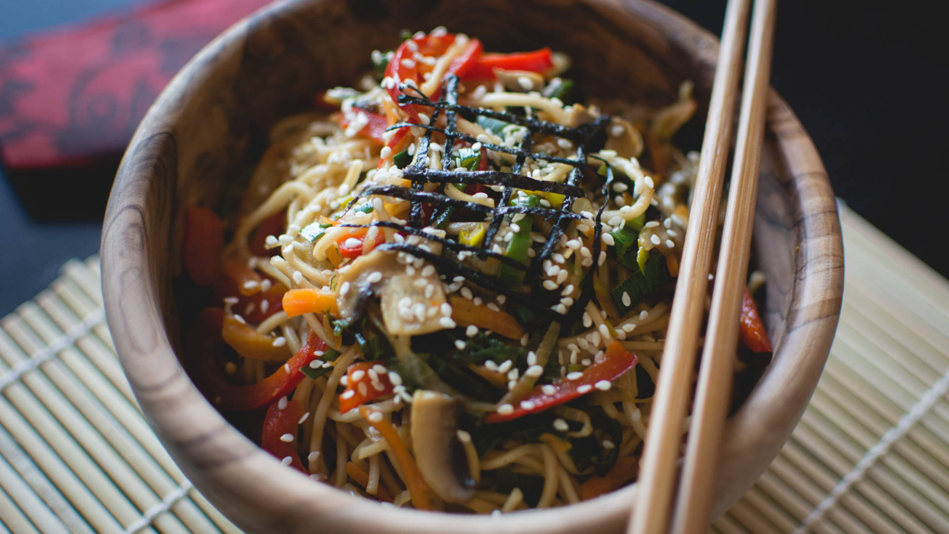 Pasta 桌布 壁紙 — 一個木碗裝滿色彩繽紛的蔬菜麵條，上面撒上芝麻和海藻醬，放在竹蓆上，旁邊放著筷子。