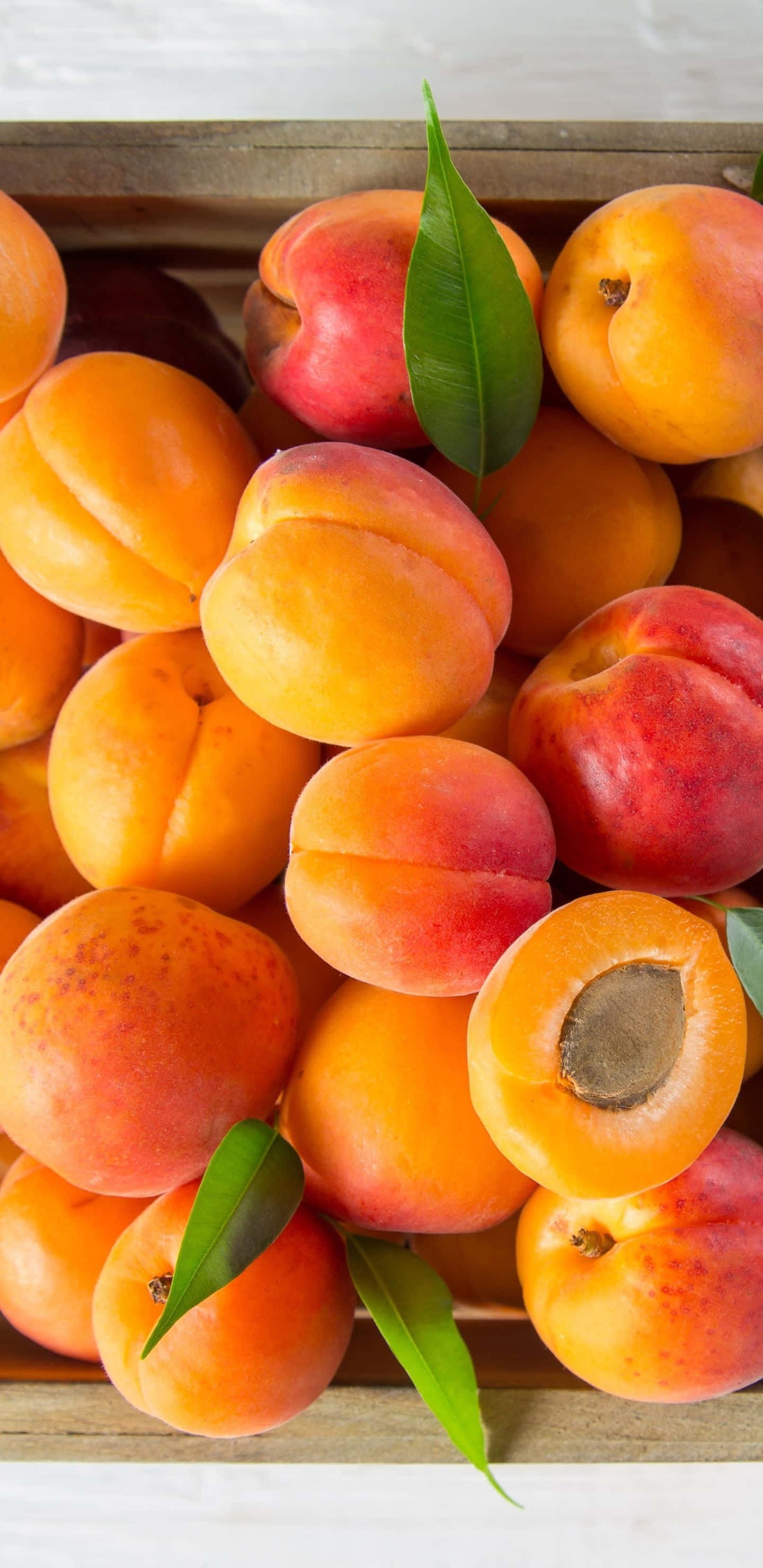 Fruit 桌布 壁紙 — 一個木箱裝滿了成熟的杏子和桃子，有些切半露出果核，並以新鮮綠葉點綴。