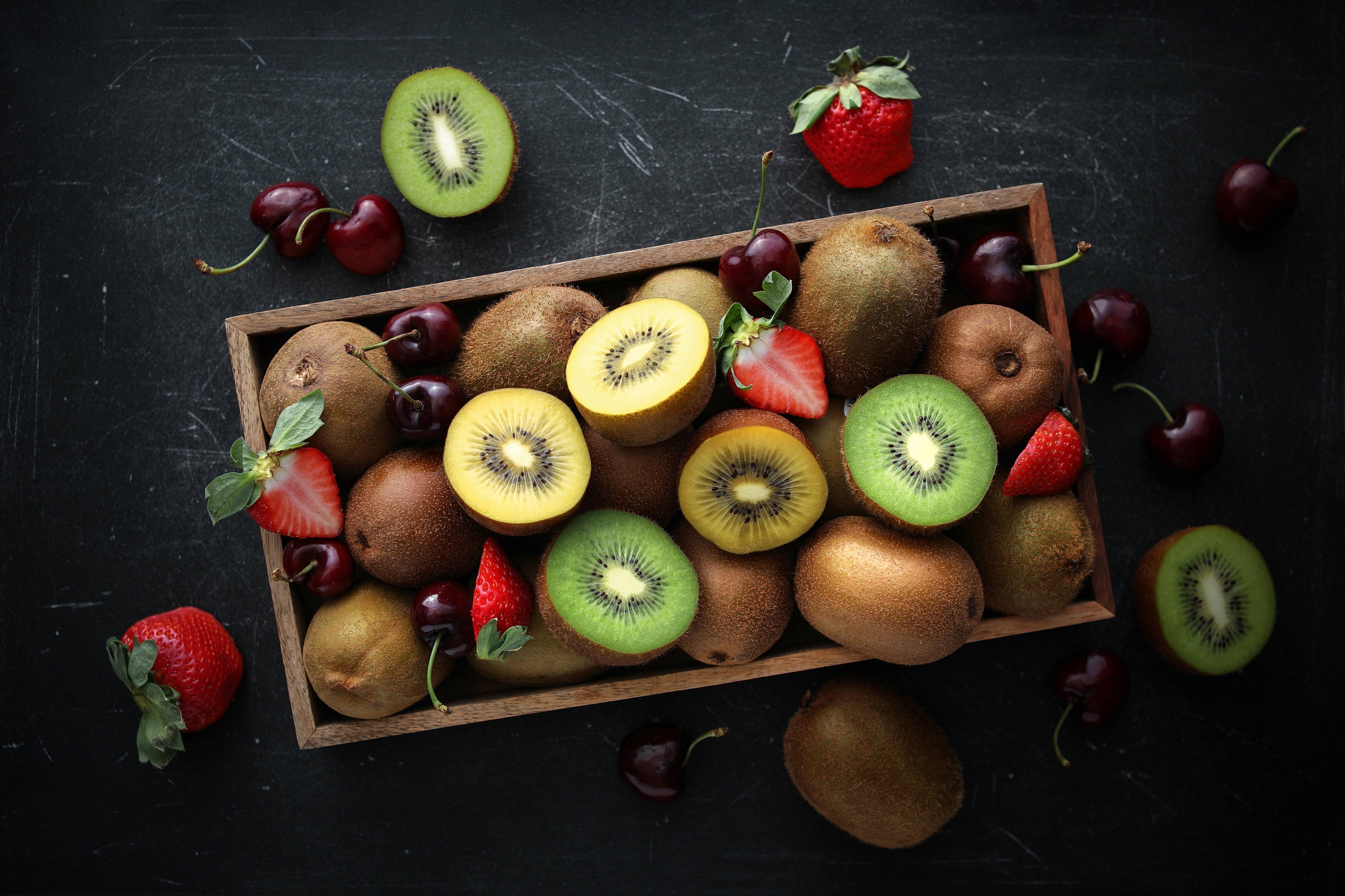 Fruits 桌布 壁紙 — 一個木箱裝滿了鮮豔的奇異果、草莓和櫻桃，排列在深色紋理表面上。