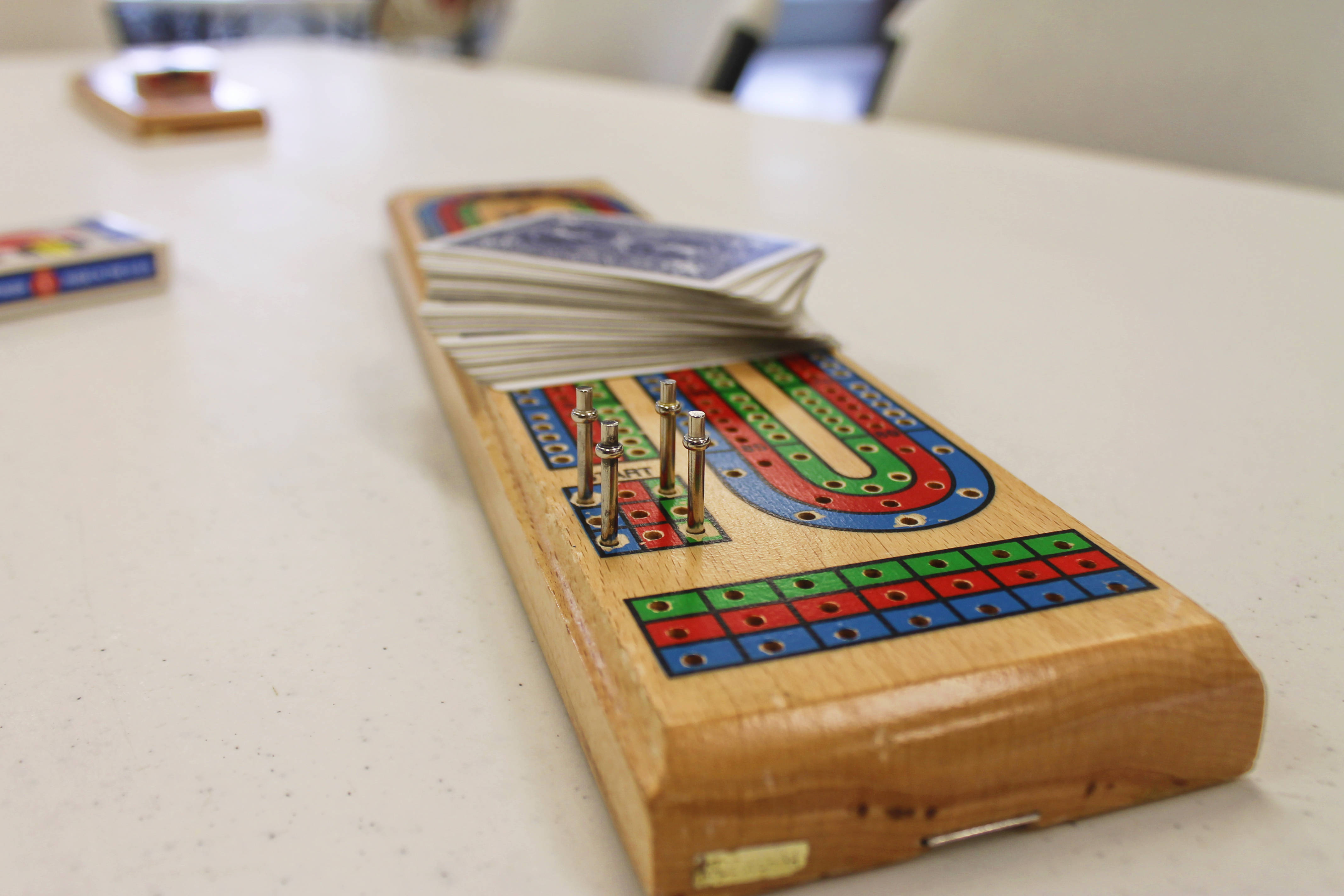 Board Game 桌布 壁紙 — 一個木製克里比奇棋盤，帶有彩色軌道和金屬釘，放在白色桌子上，附近有一副紙牌。