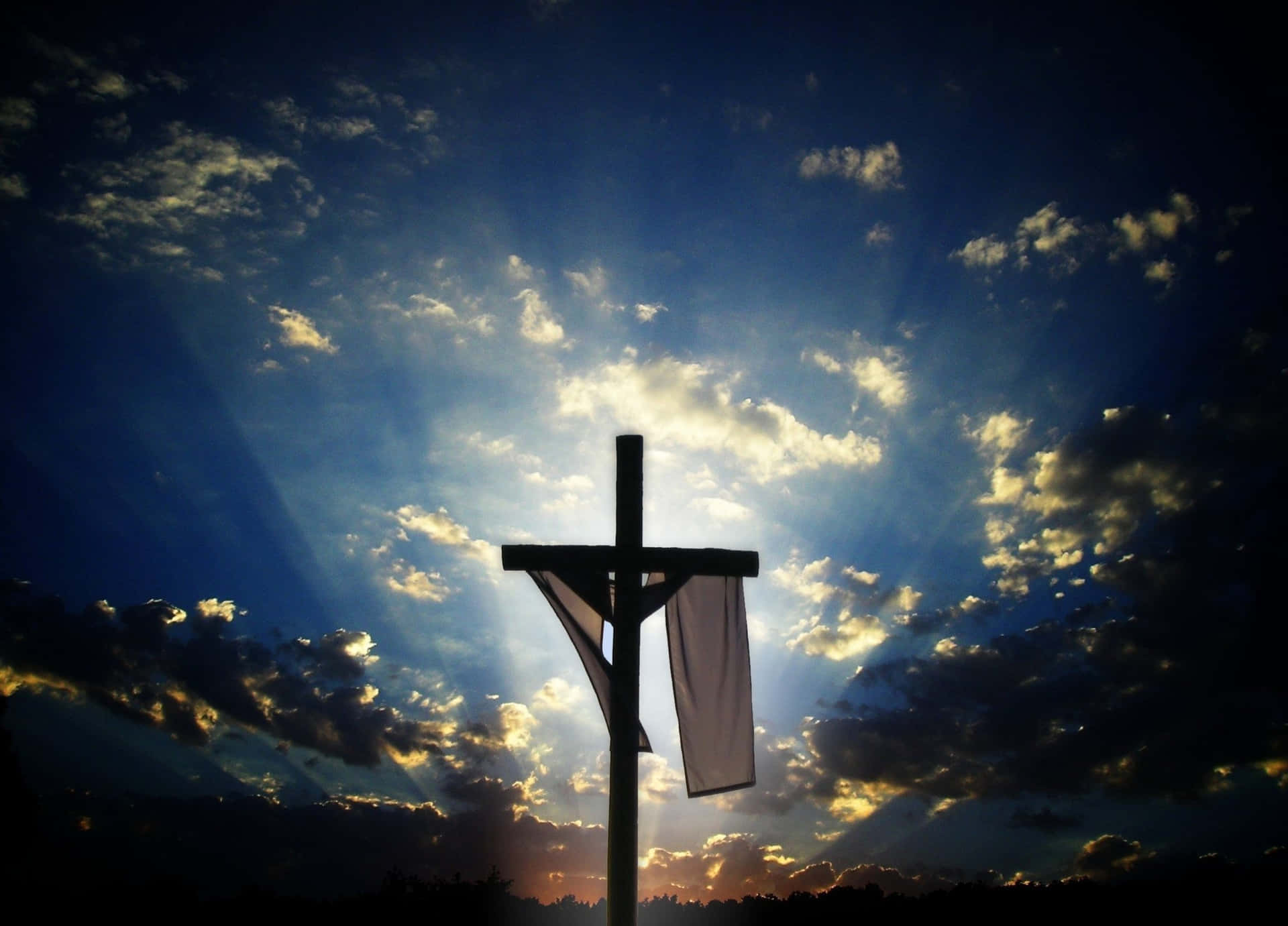 Easter 桌布 壁紙 — 一個木製十字架在日落時分，以戲劇性的天空為背景，金色雲層中透出耀眼陽光，形成剪影。