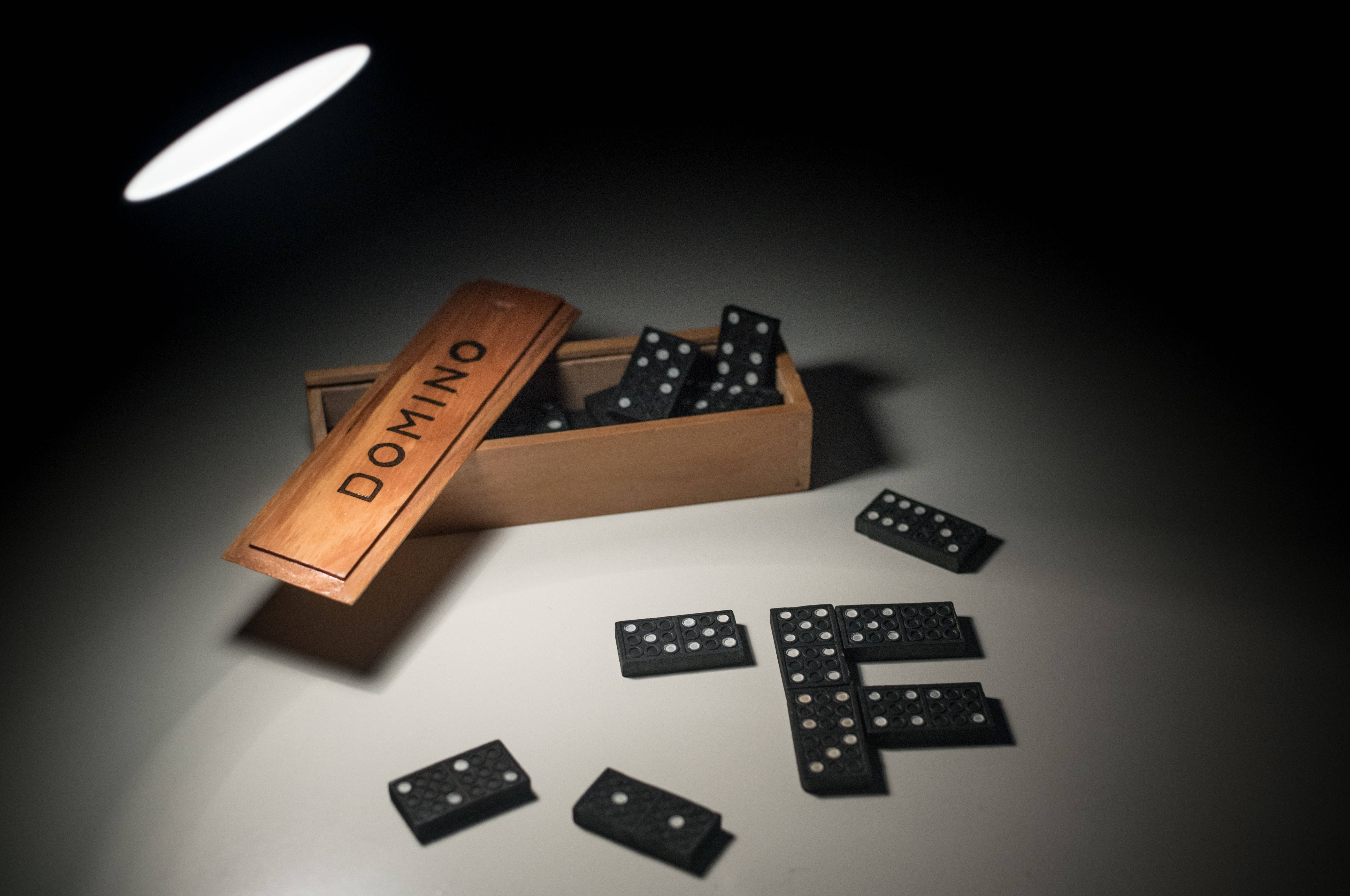 dominos 桌布 壁紙 — 一個木製多米諾骨牌盒，內有黑色多米諾骨牌，在戲劇性的聚光燈下灑落在純白表面上。