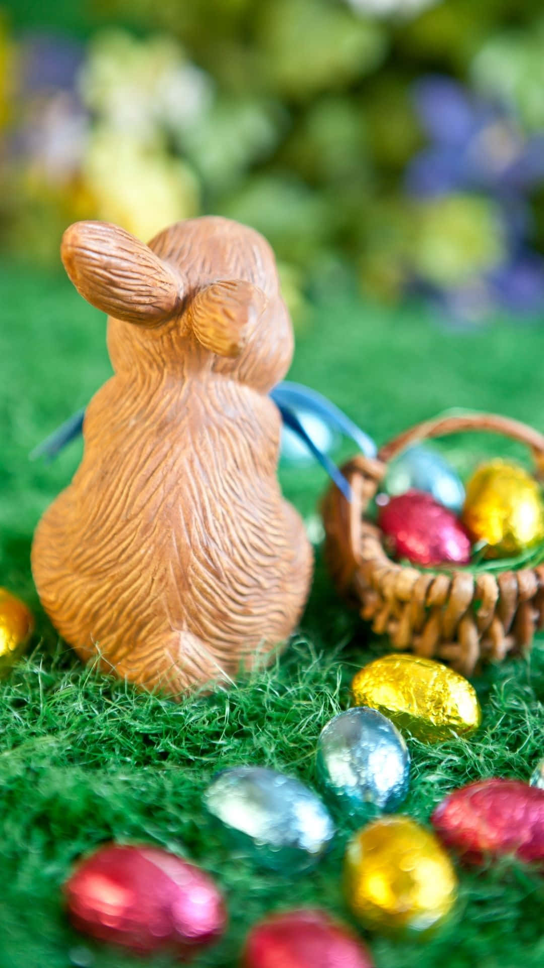 Easter Bunny 桌布 壁紙 — 一個木製復活節兔公仔坐在綠色草地上，旁邊有一個柳條籃和用錫箔紙包裹的巧克力蛋。
