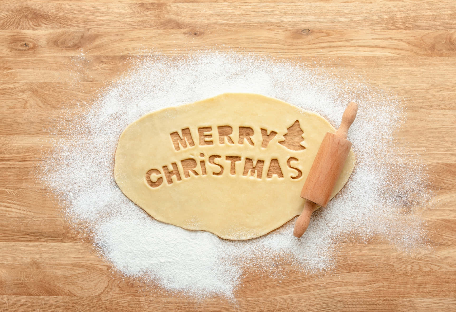 Merry Christmas Dough & Flour Image 壁紙 — 一個木製擀麵棍放在一個扁平麵糰圈旁邊，麵糰上印有「MERRY CHRISTMAS」和一棵樹的圖案，在木質表面上用麵粉呈現。