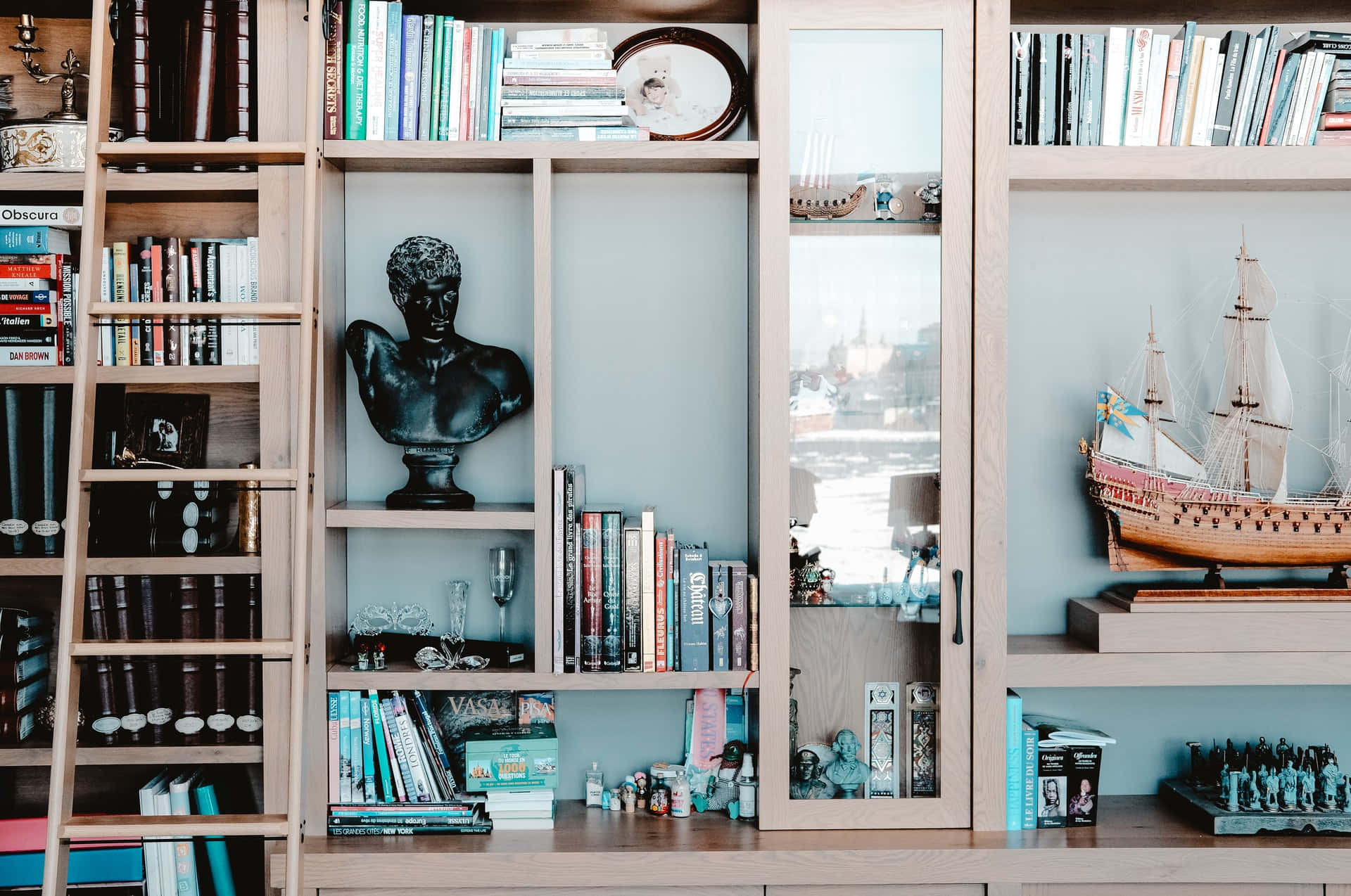 Bookshelf 桌布 壁紙 — 一個木製書架，裝滿書籍、雕塑和模型船，透過玻璃門反射出城市景觀。